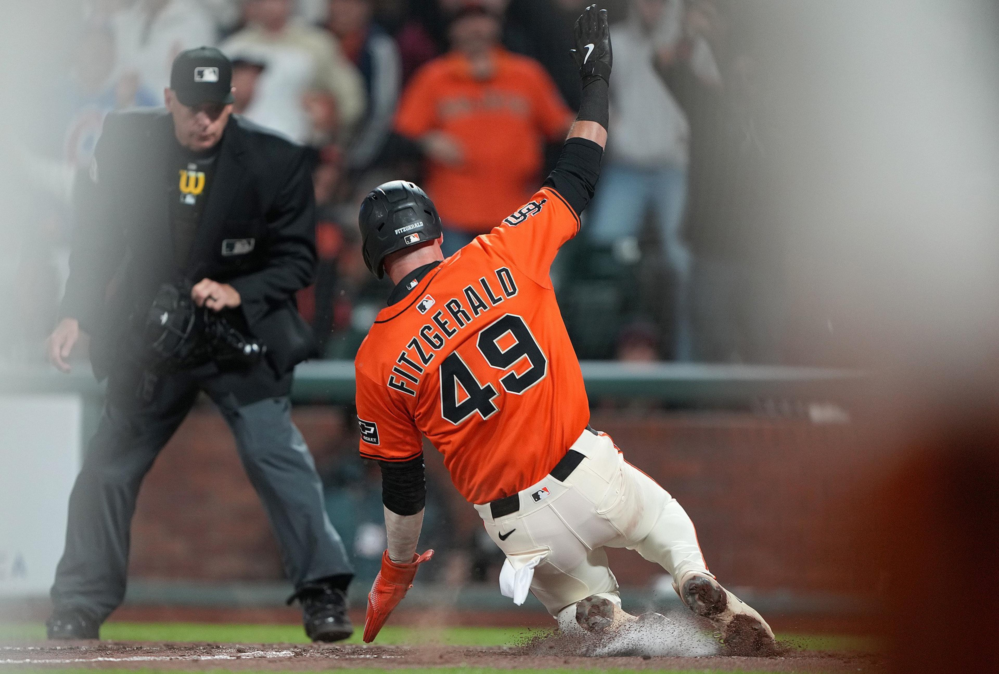 A baseball player in an orange jersey with the number 49 slides into a base. An umpire in black watches closely, and the scene is blurred around the edges.