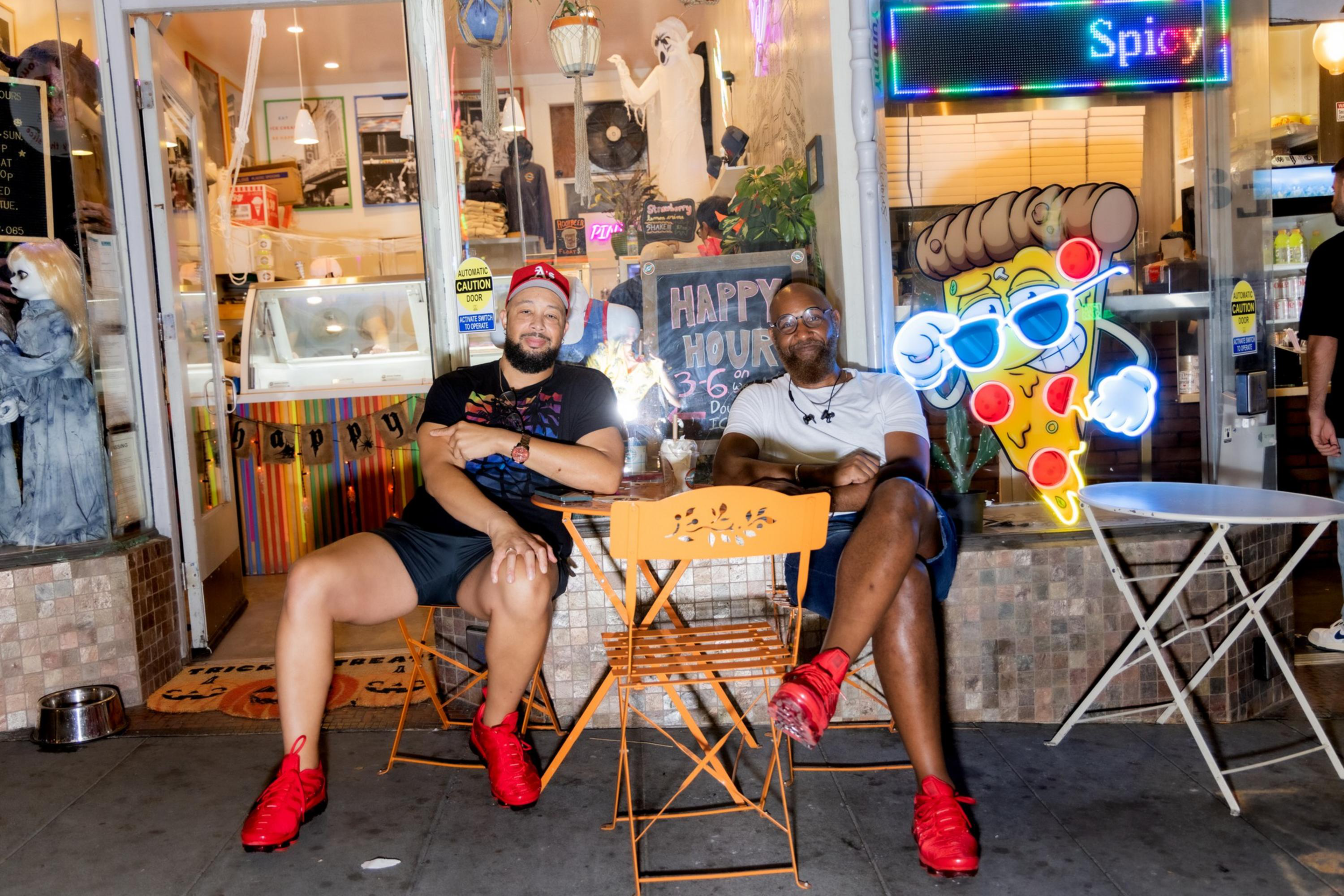 Two men wearing red shoes sit on orange chairs outside a shop with a smiling pizza slice sign and a happy hour chalkboard.
