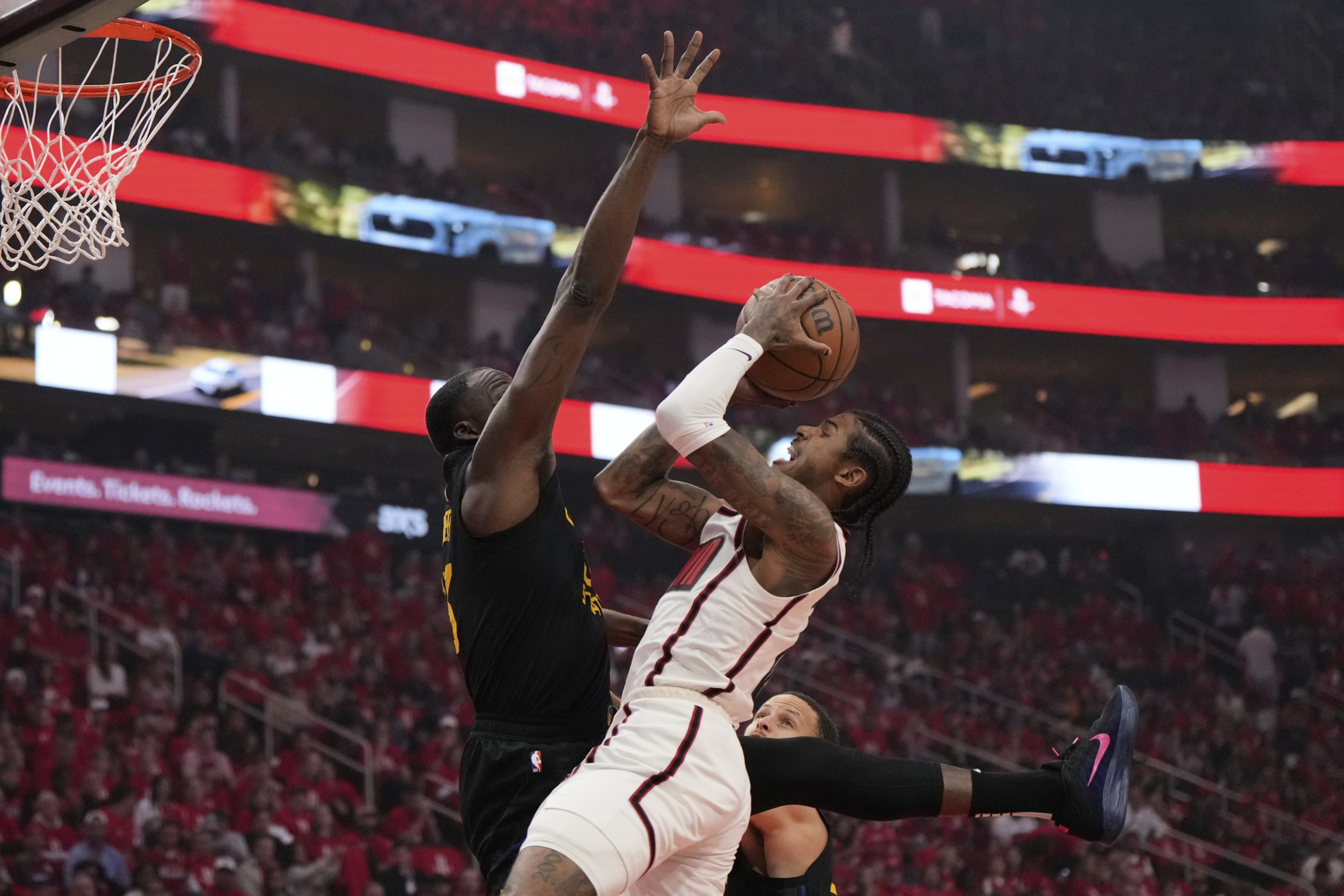 A basketball player in a white jersey jumps to shoot the ball, while a defender in a black jersey attempts to block him. The background shows a crowded arena.