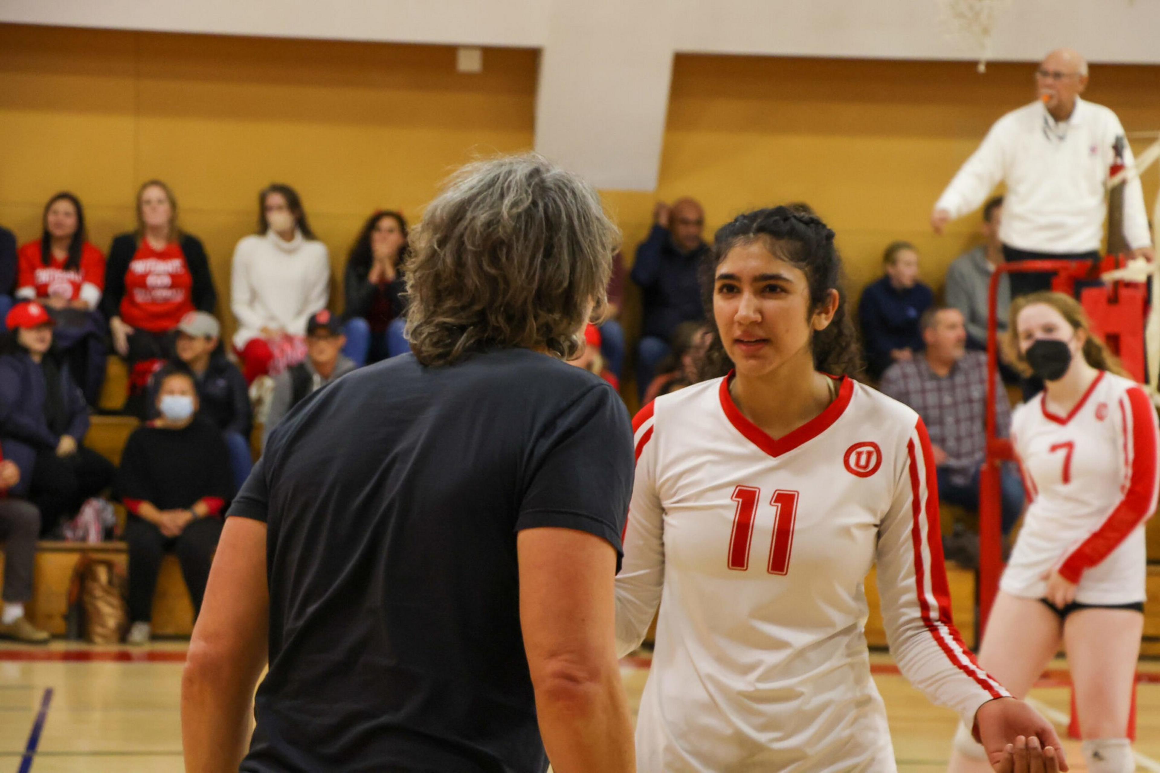 A volleyball player in a white and red jersey numbered 11 listens intently to a coach in a black shirt during a game, spectators watch in the background.