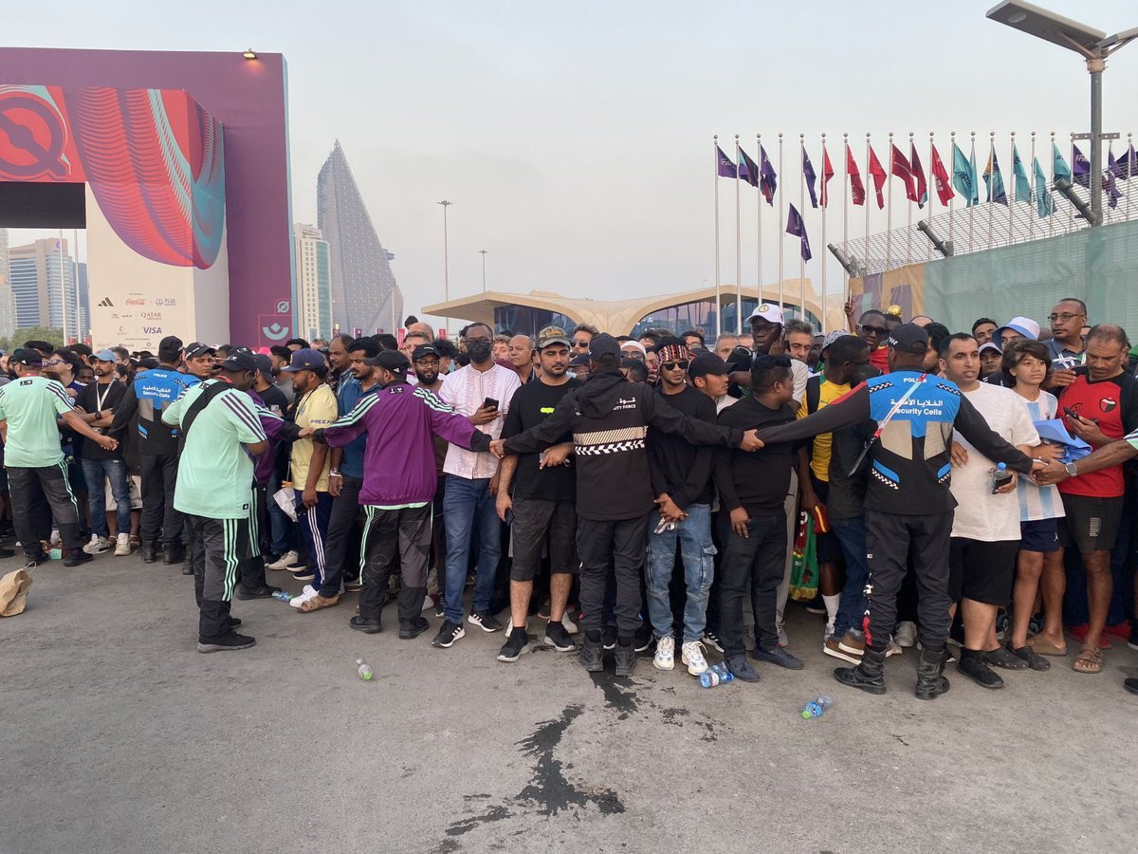 A group of security guards forms a human barrier, holding hands to manage a crowd of people gathered near a large event entrance and flags.