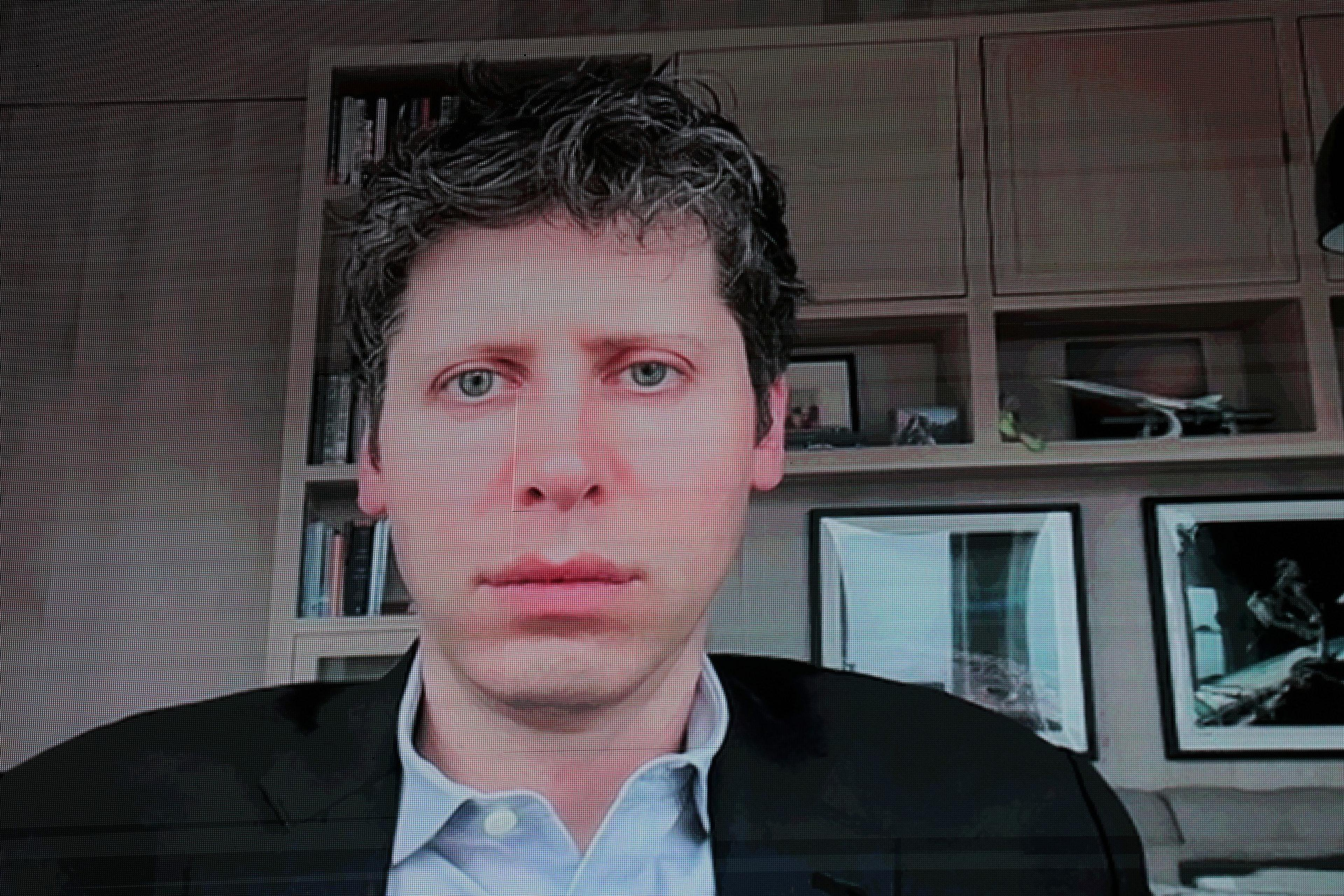 A man with curly dark hair and blue eyes wears a dark blazer over a light blue shirt, sitting in front of a bookshelf and framed pictures.
