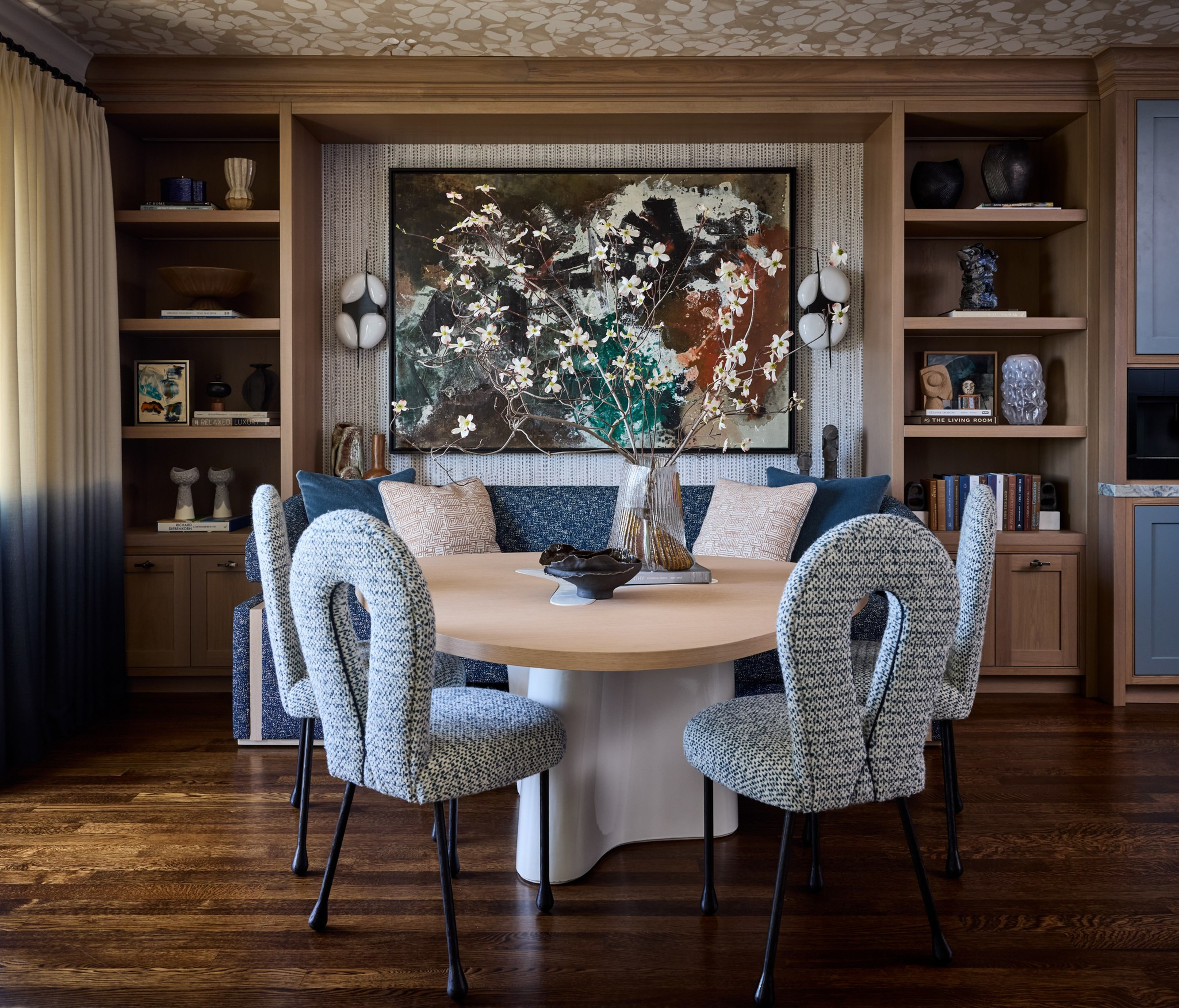 A round wooden table with a floral centerpiece is surrounded by four patterned chairs, set in a cozy room with built-in wooden shelves and abstract art.