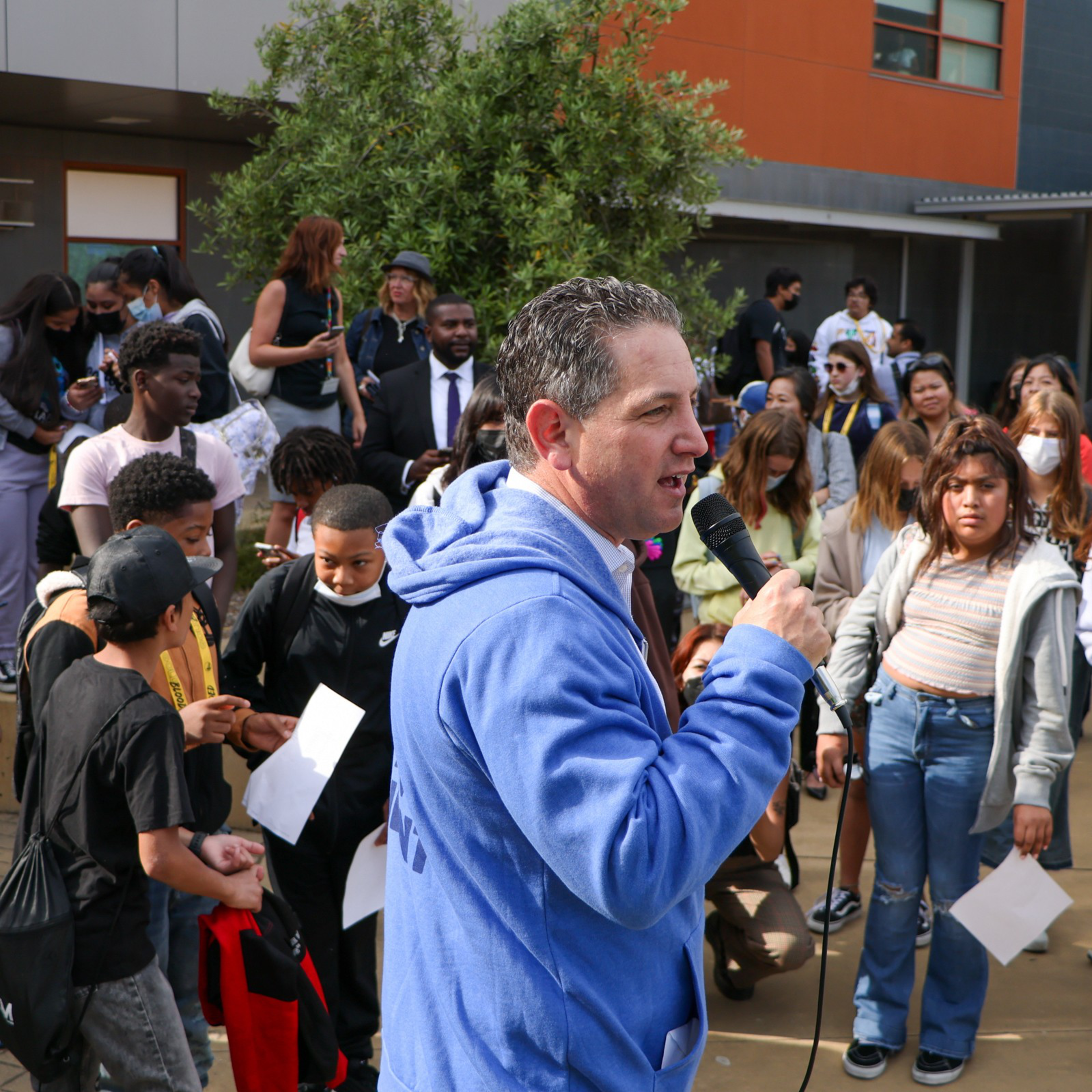 A man in a blue hoodie speaks into a microphone in front of a diverse crowd, including children and adults, some of whom are holding papers and wearing backpacks.