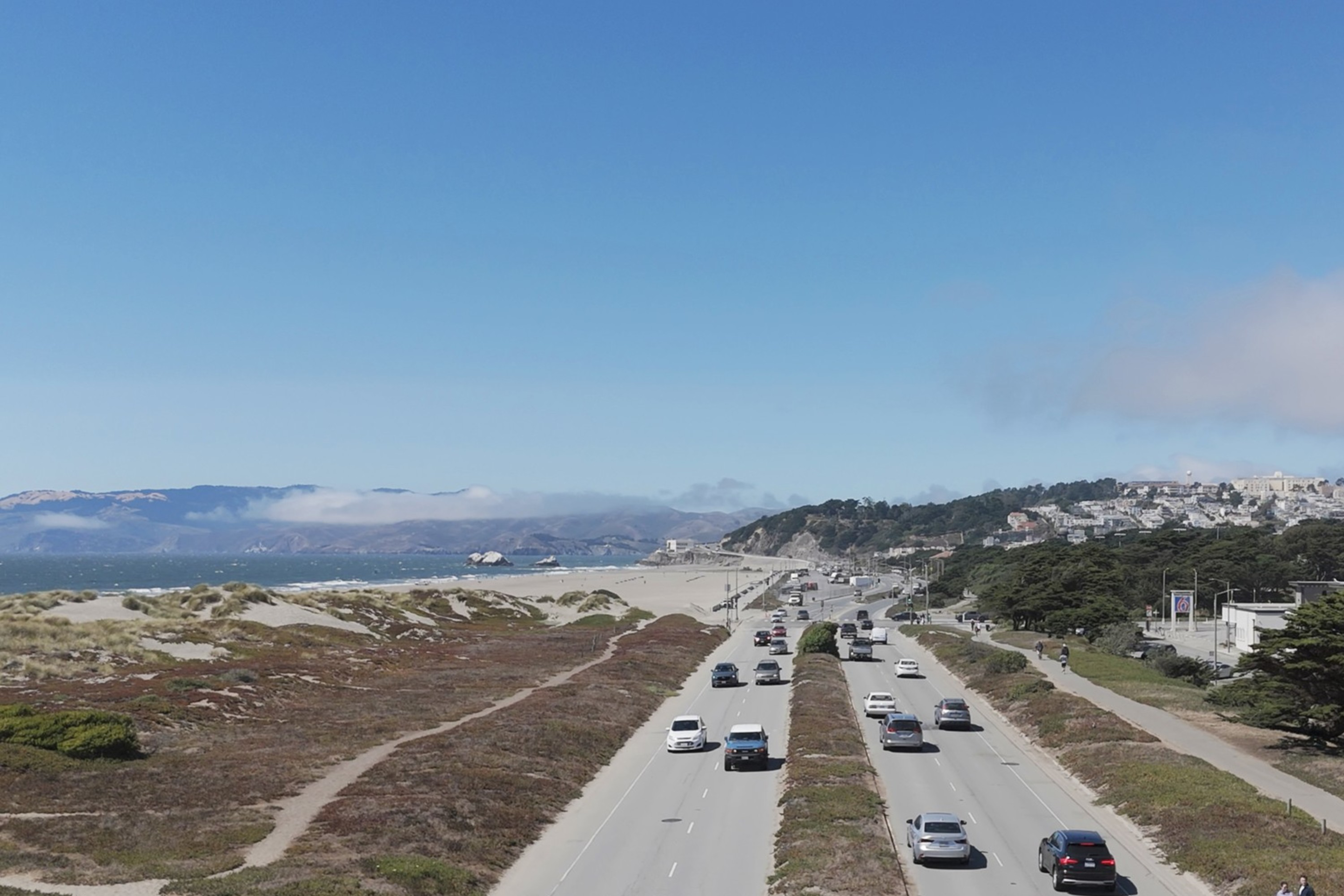 The image shows a road with cars driving between dunes and greenery. A clear blue sky and distant hills meet a cityscape, featuring modern buildings and a large windmill.