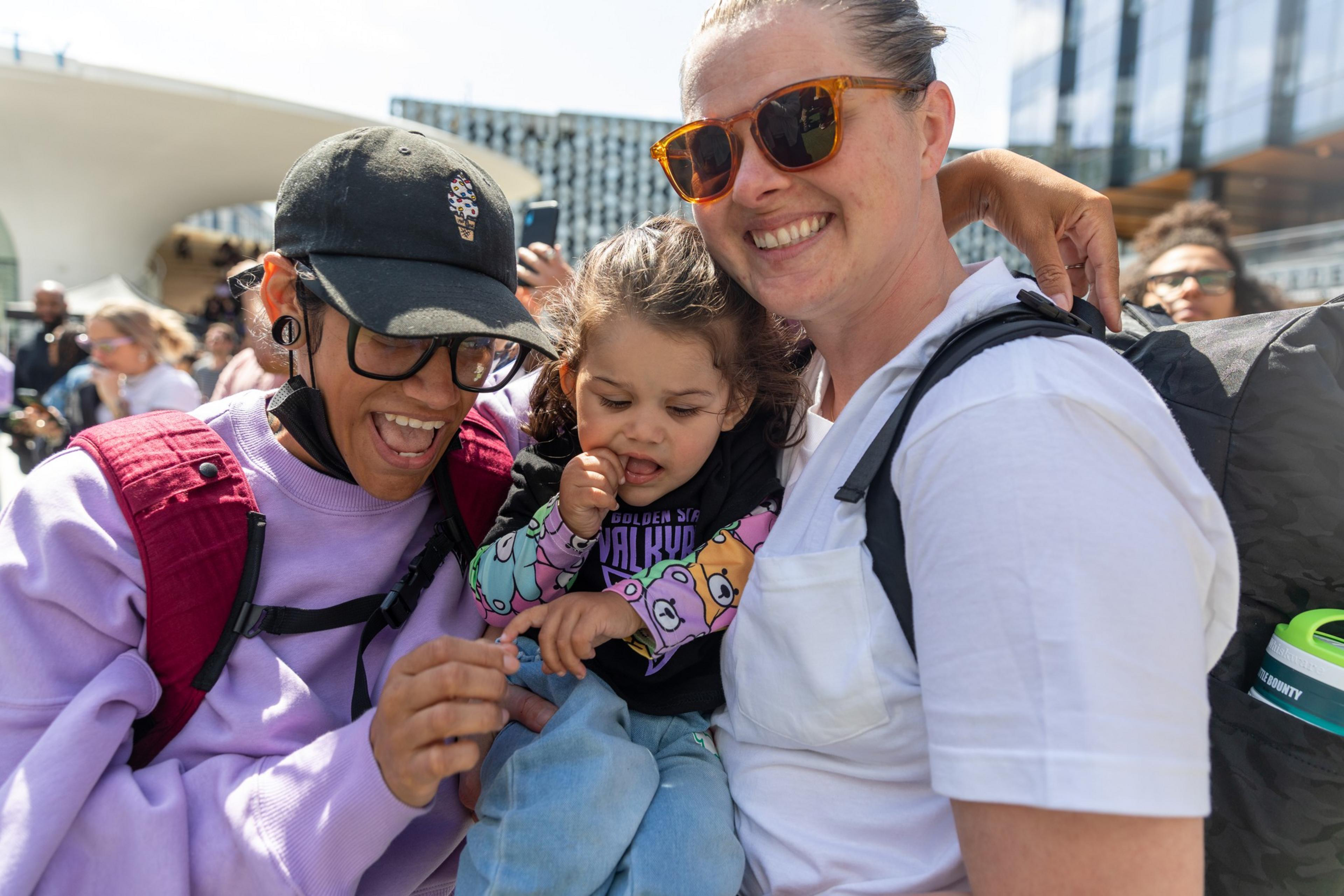 Two women and a child, smiling and together outdoors, surrounded by others in a bright, lively setting. One woman wears sunglasses; the other, a baseball cap.