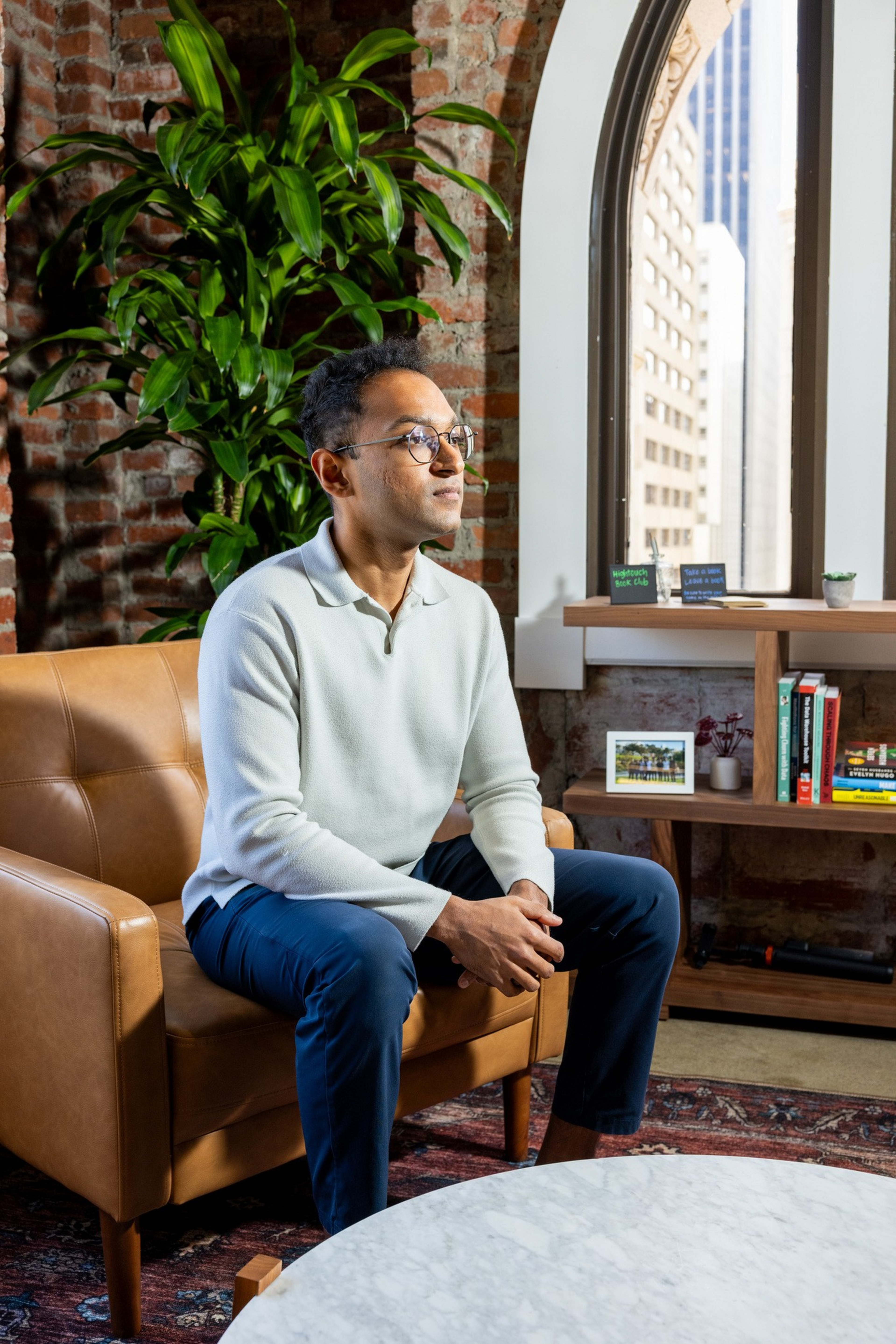 A man wearing glasses, a white long-sleeve shirt, and blue pants sits on a brown leather chair in a room with a brick wall and large window.