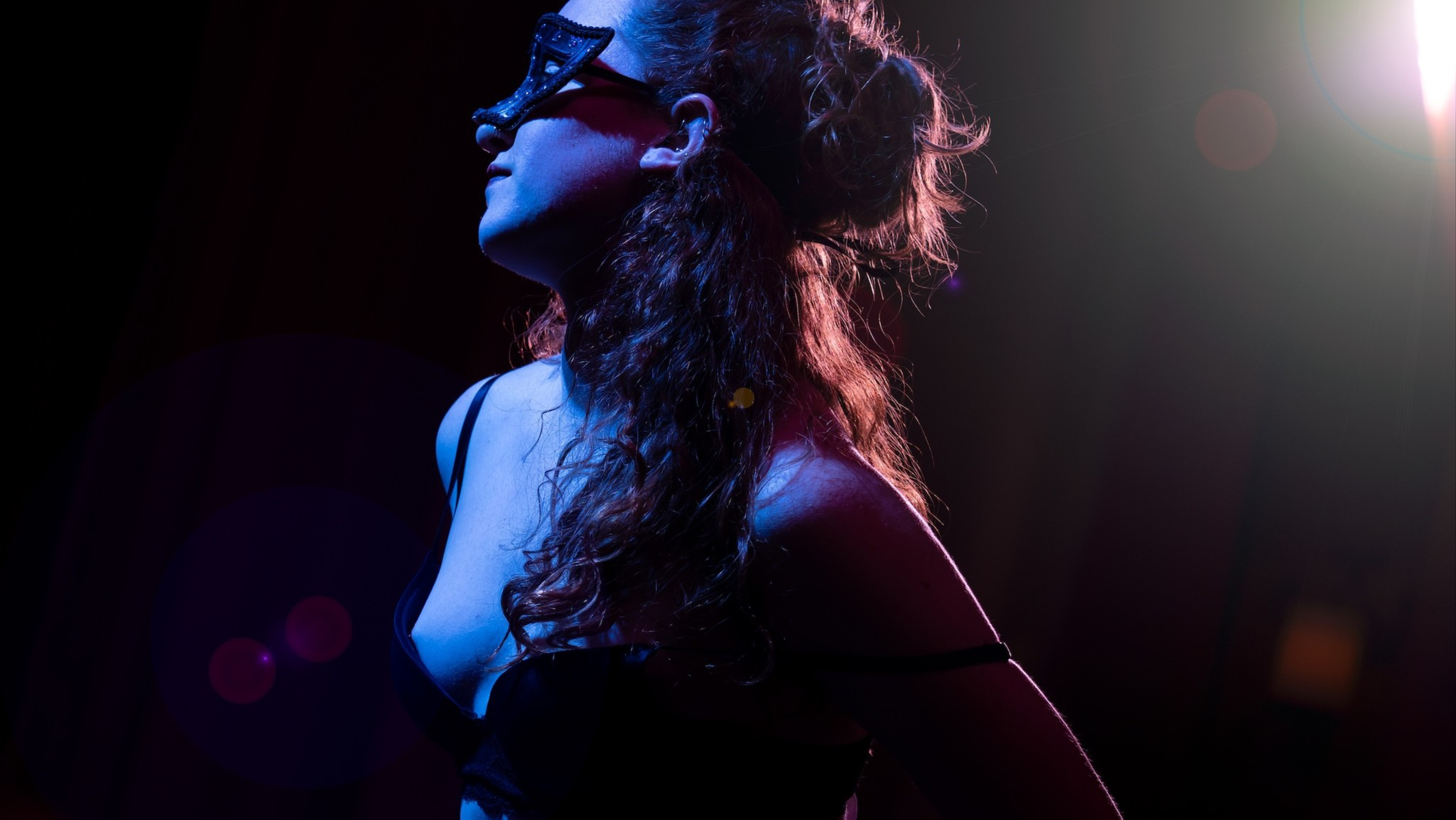 A woman with curly hair in a black strapless top and black mask looks upward, illuminated by blue and purple stage lights against a dark background.