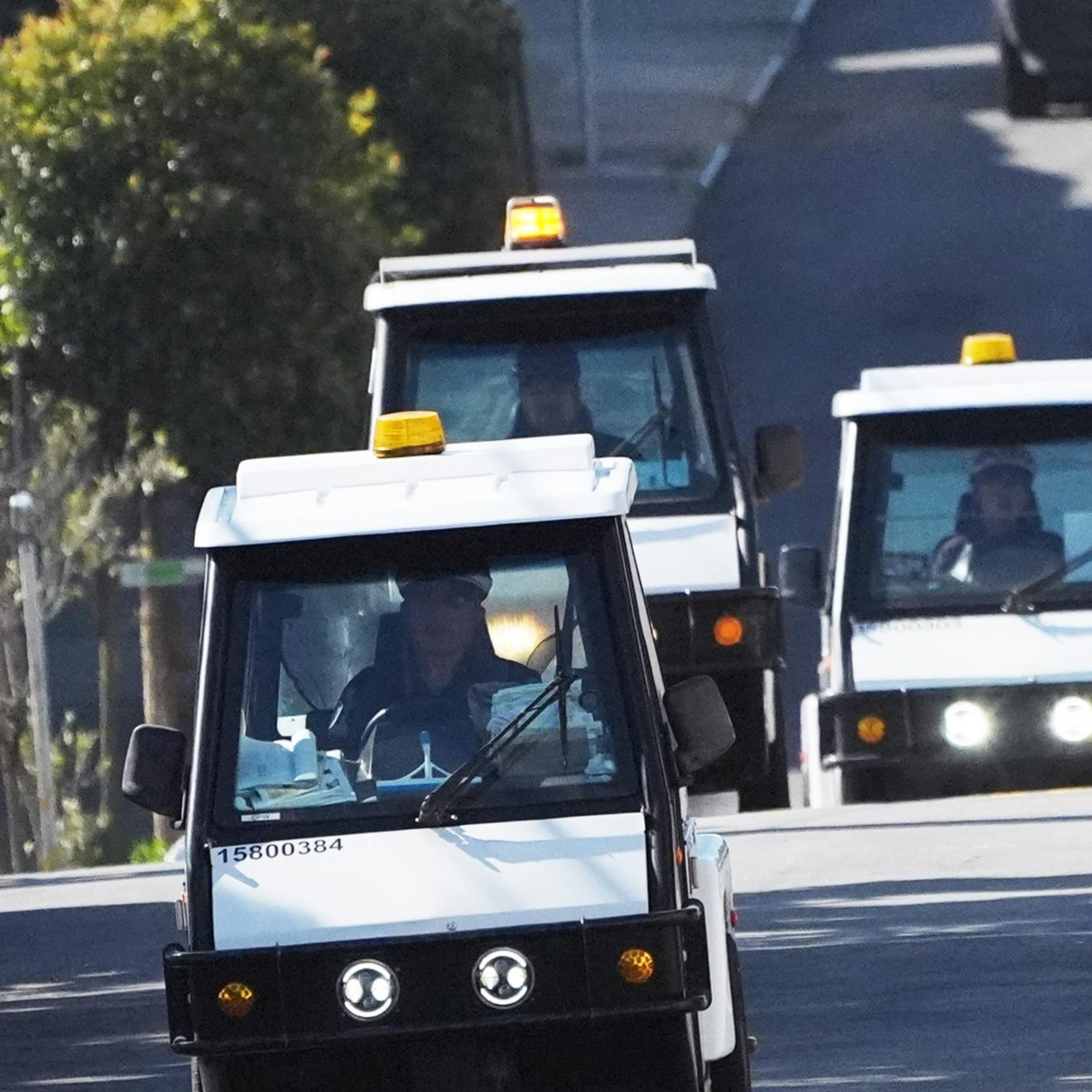 A convoy of small white utility vehicles, each with an amber light on top, drives down a road.