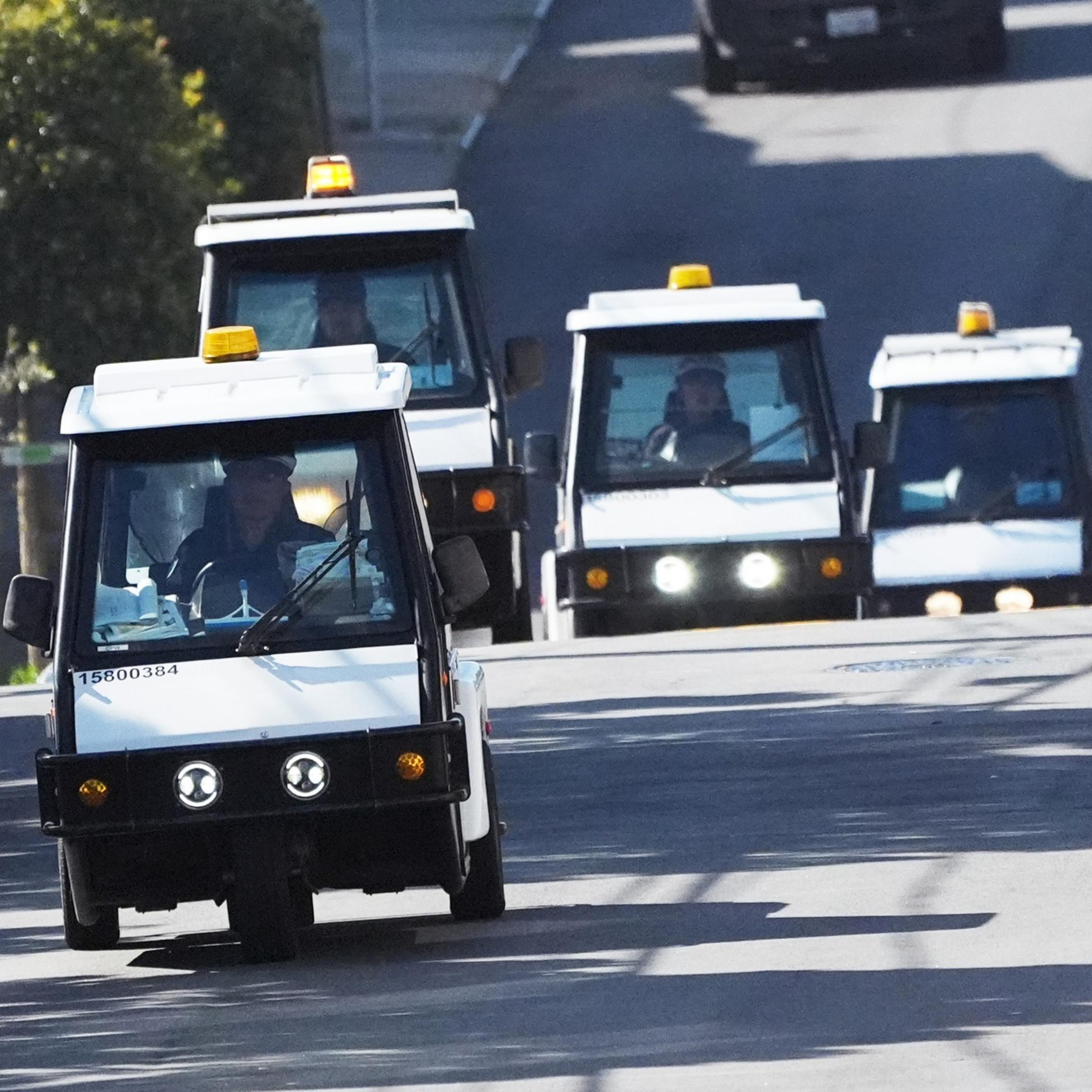 A convoy of small white utility vehicles, each with an amber light on top, drives down a road.