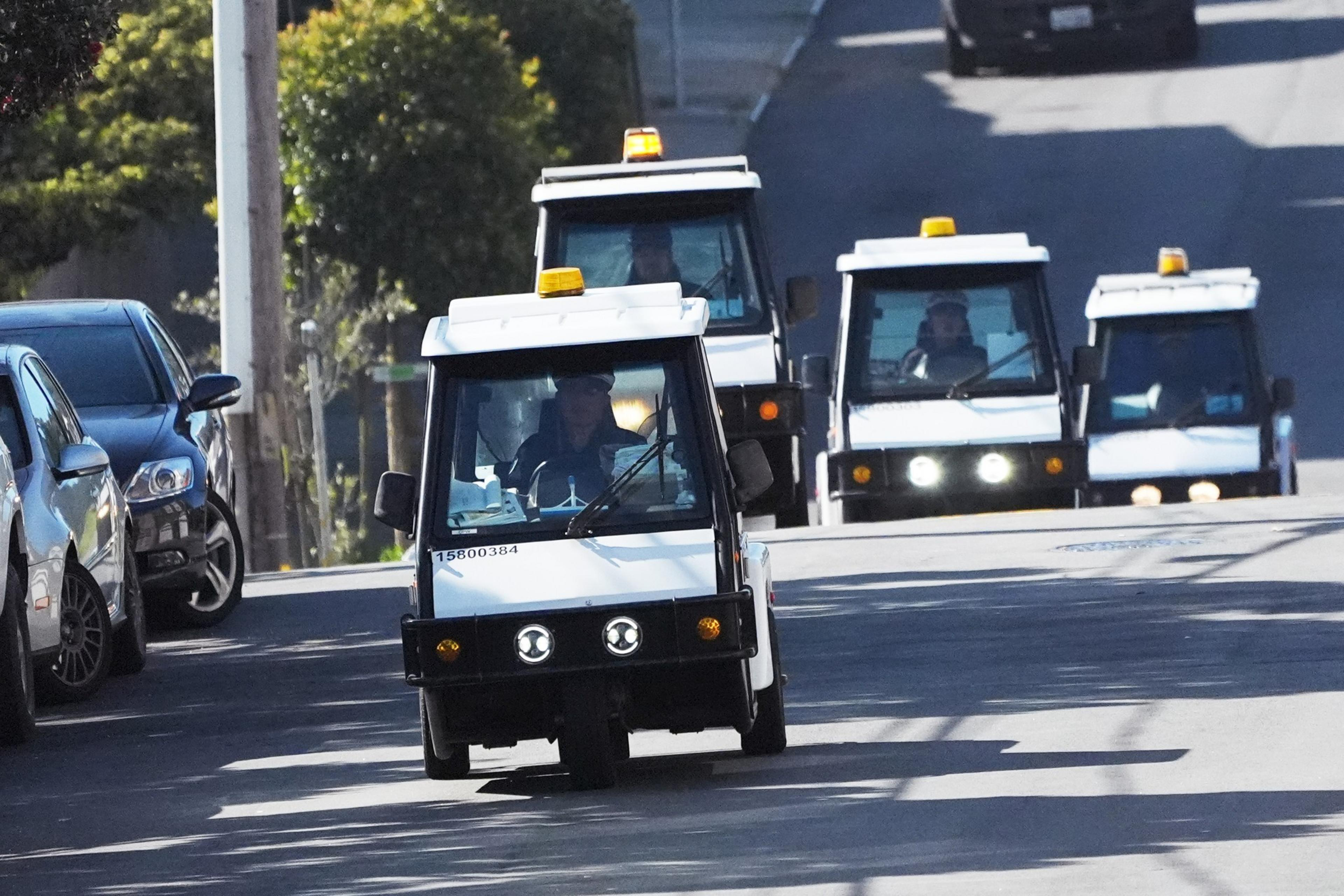 A convoy of small white utility vehicles, each with an amber light on top, drives down a road.