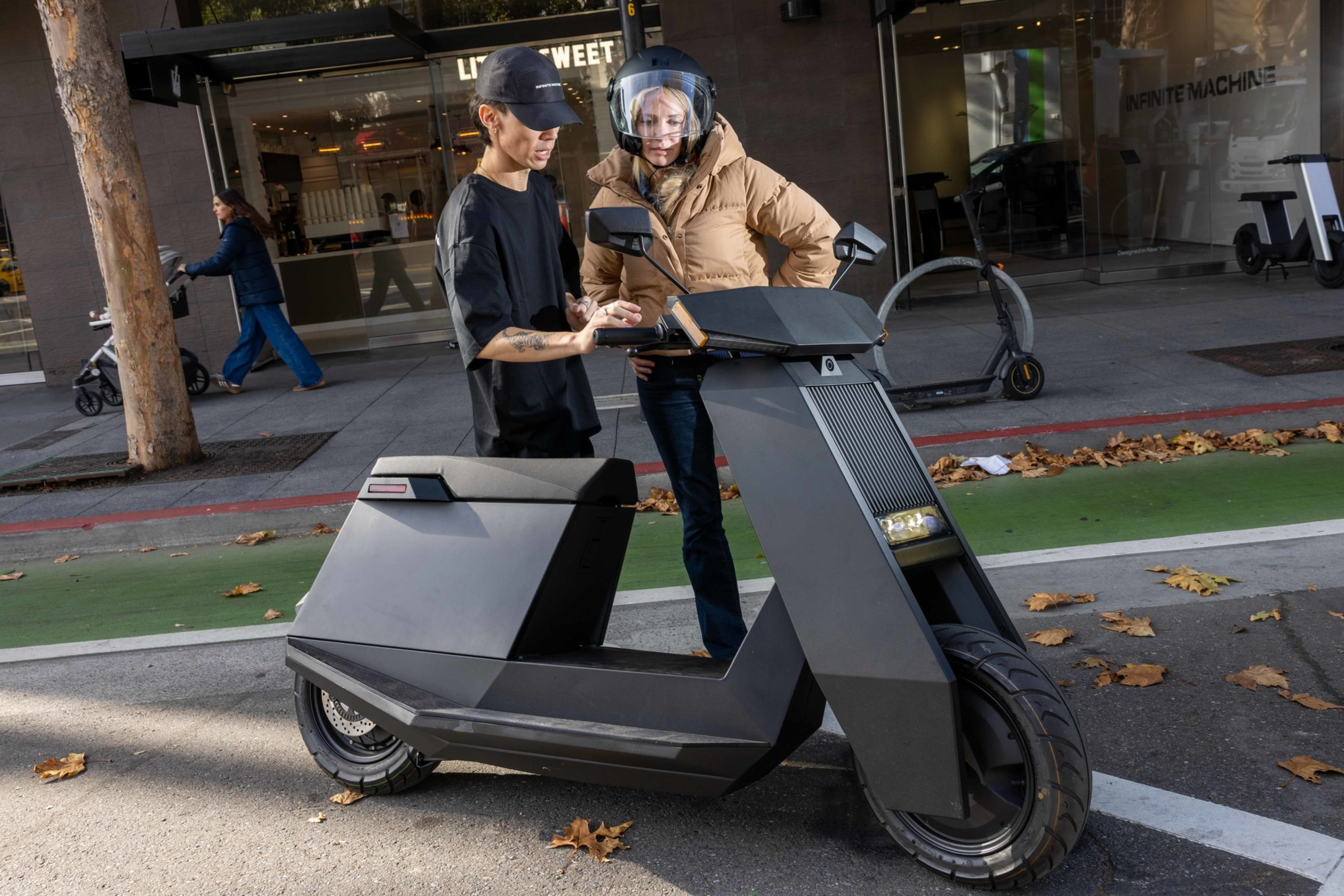 Two women stand next to a sleek black electric scooter on a city street, with one woman showing something on a device to the other.
