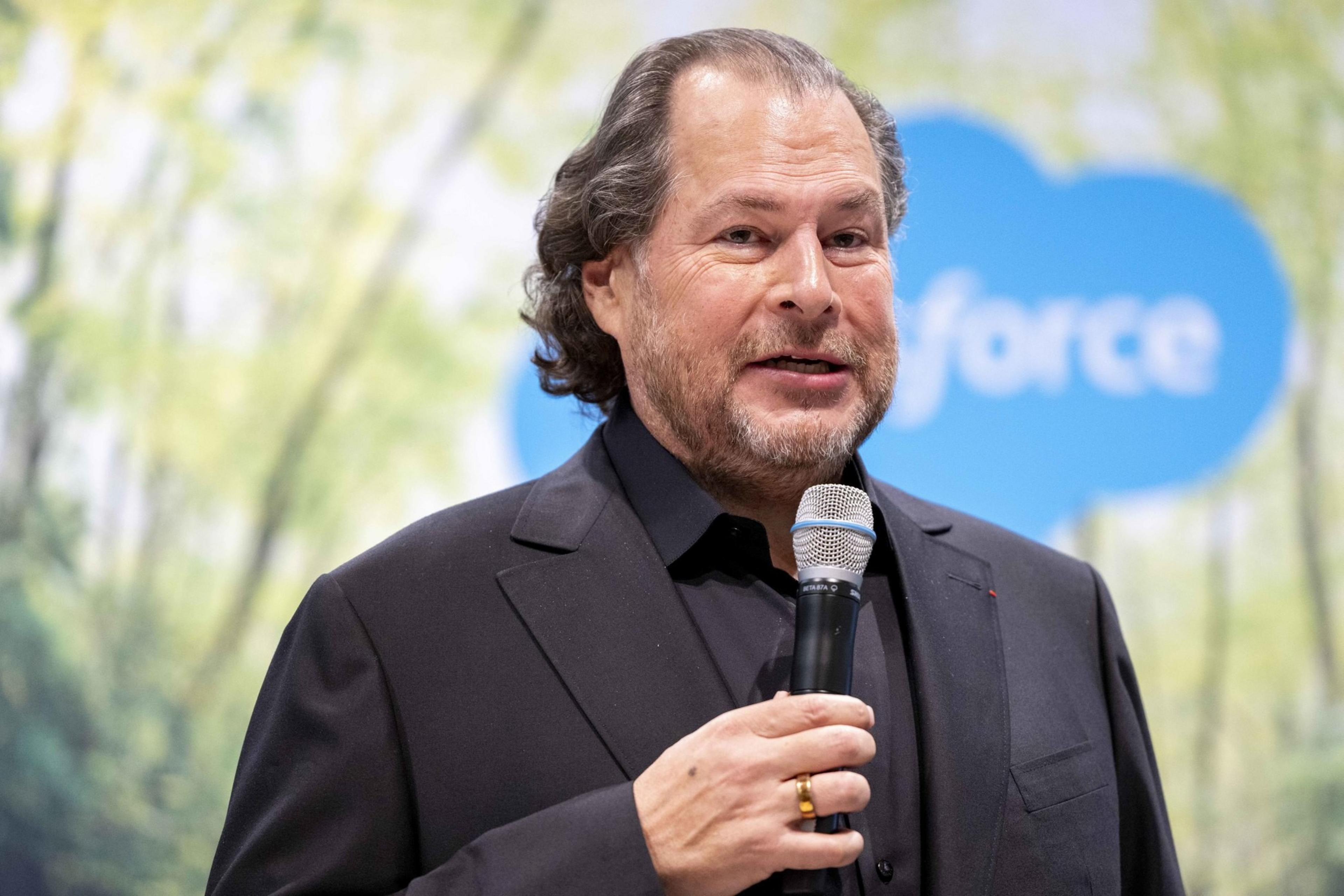 A man in a black suit holds a microphone, speaking against a blurred background with a blue logo partially visible. He has short, wavy hair and is smiling slightly.