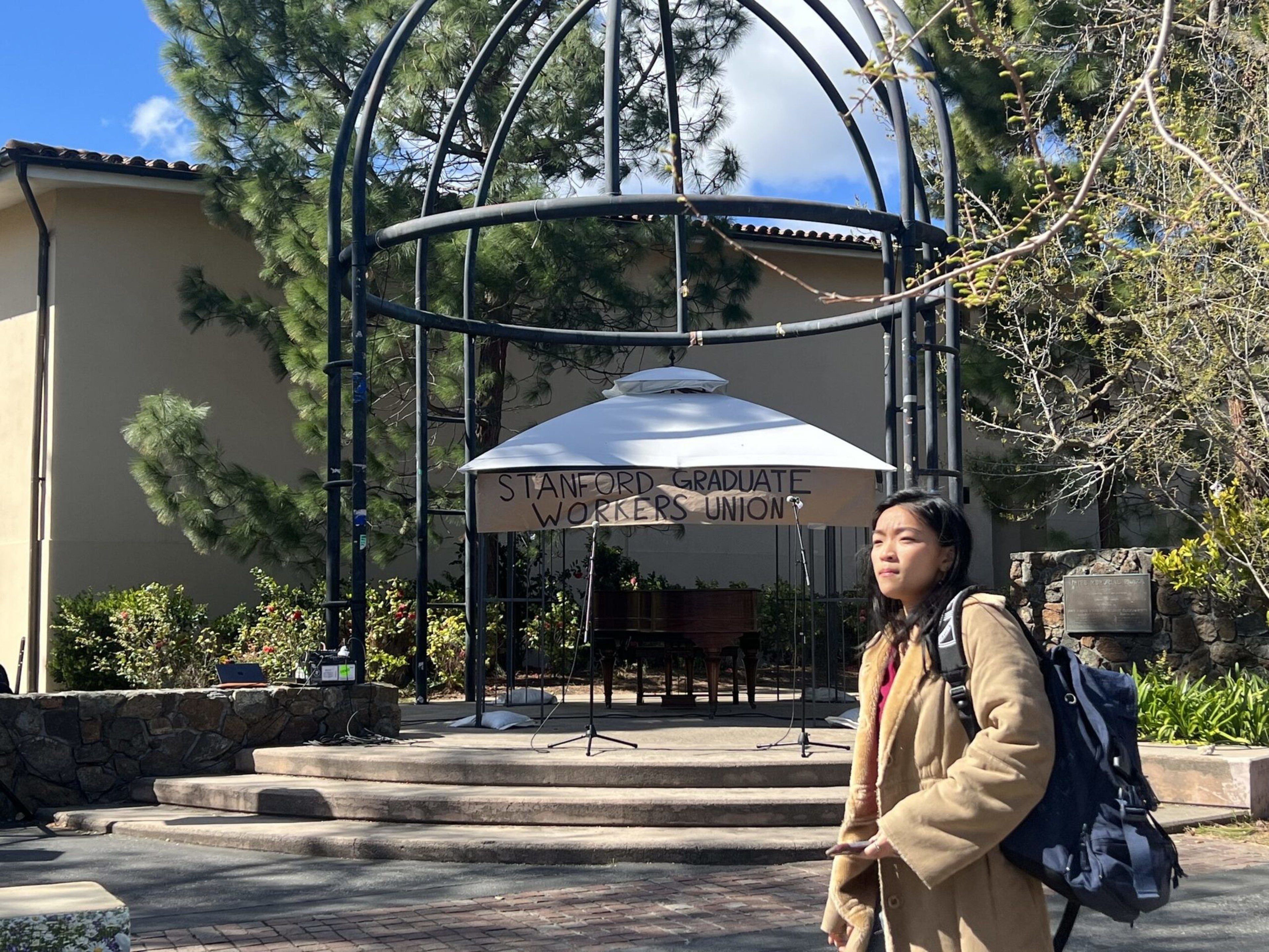 A woman with a backpack walks by an outdoor stage with a sign that reads "Stanford Graduate Workers Union" under a metal gazebo.