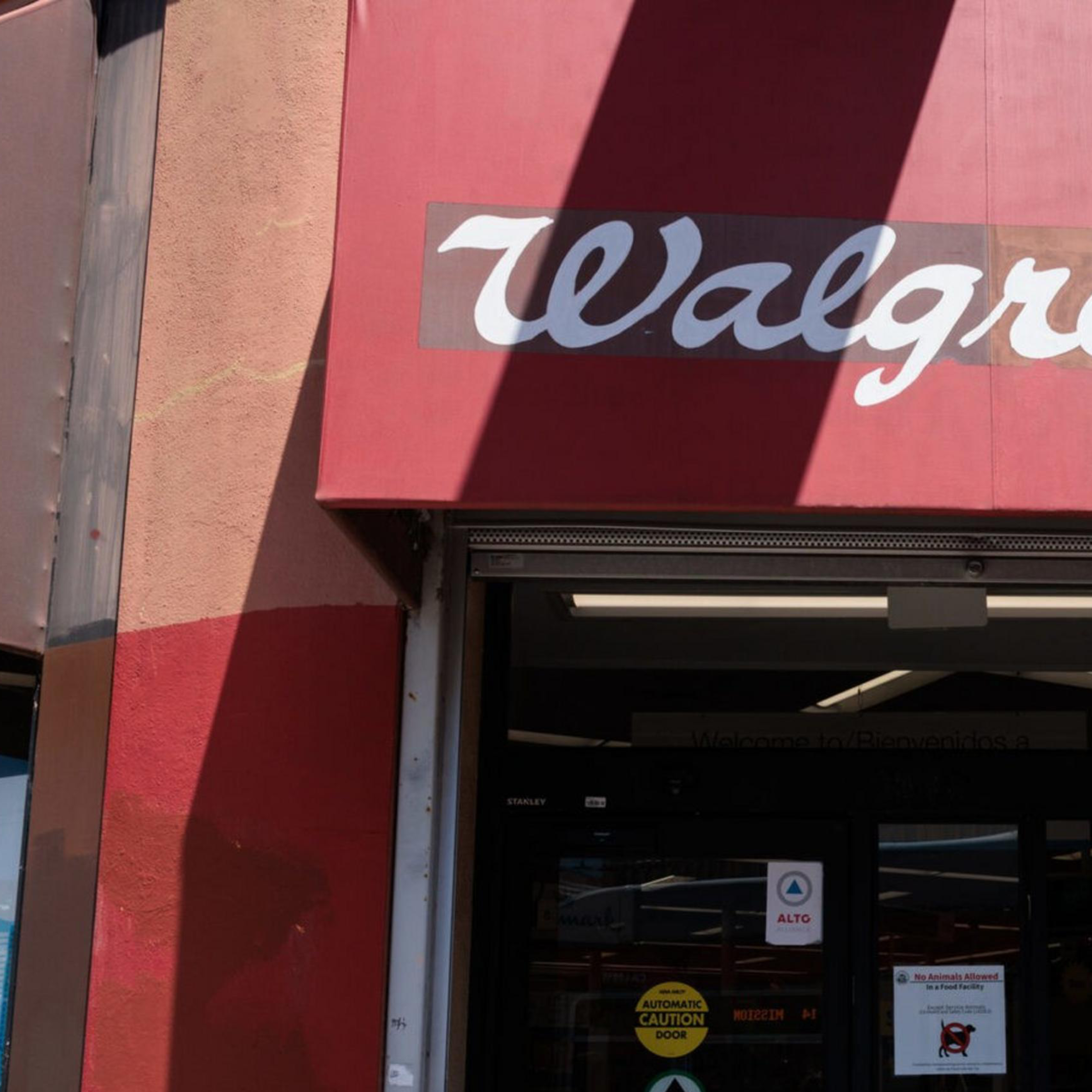 A person is exiting a Walgreens store, wearing a dark hoodie and carrying a drink. The storefront features a red awning with the Walgreens logo.