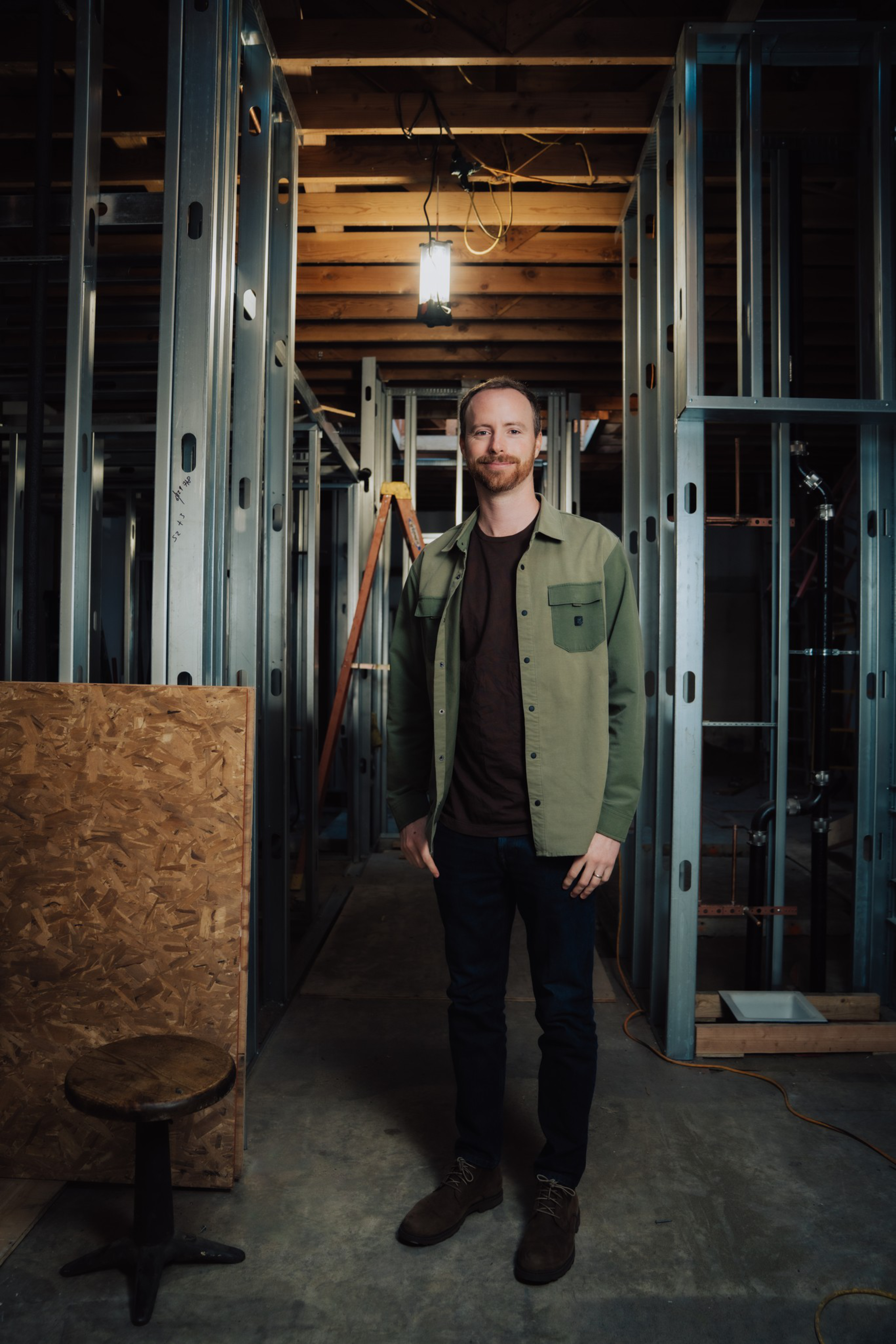 A man with a beard stands smiling inside a dimly lit construction site framed by metal studs, wearing a green jacket, dark pants, and brown shoes.