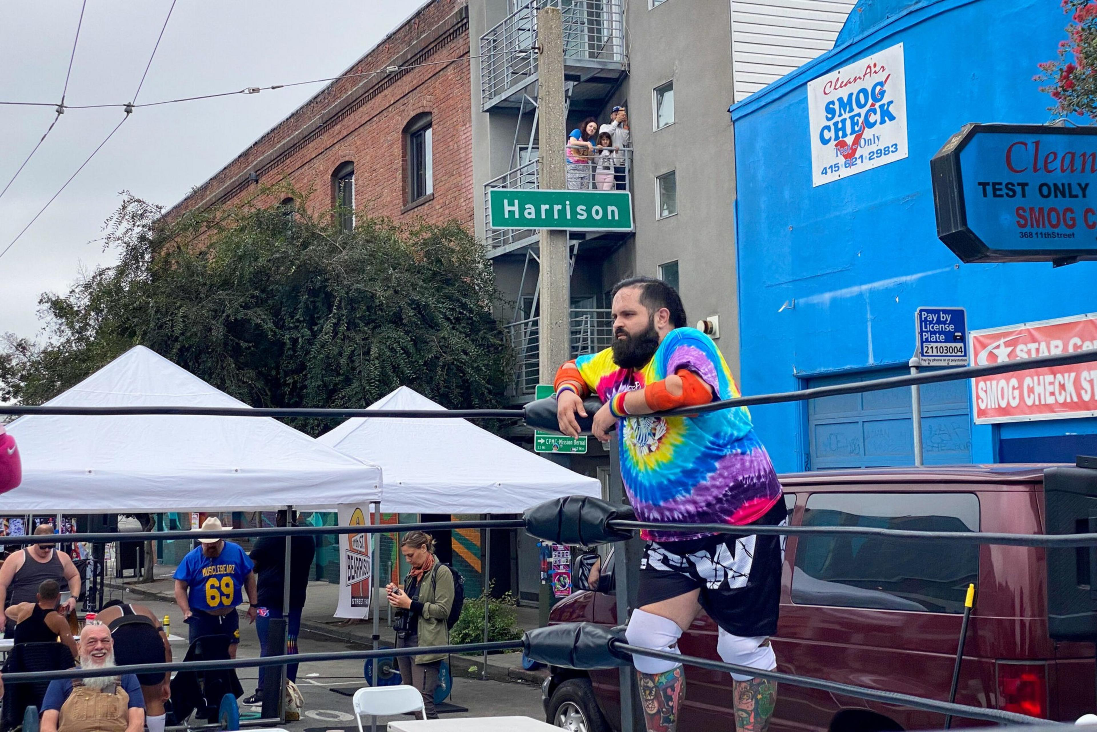 A man in a colorful tie-dye shirt and wrestling gear leans on the ropes of a wrestling ring set up outdoors near a street sign reading "Harrison."
