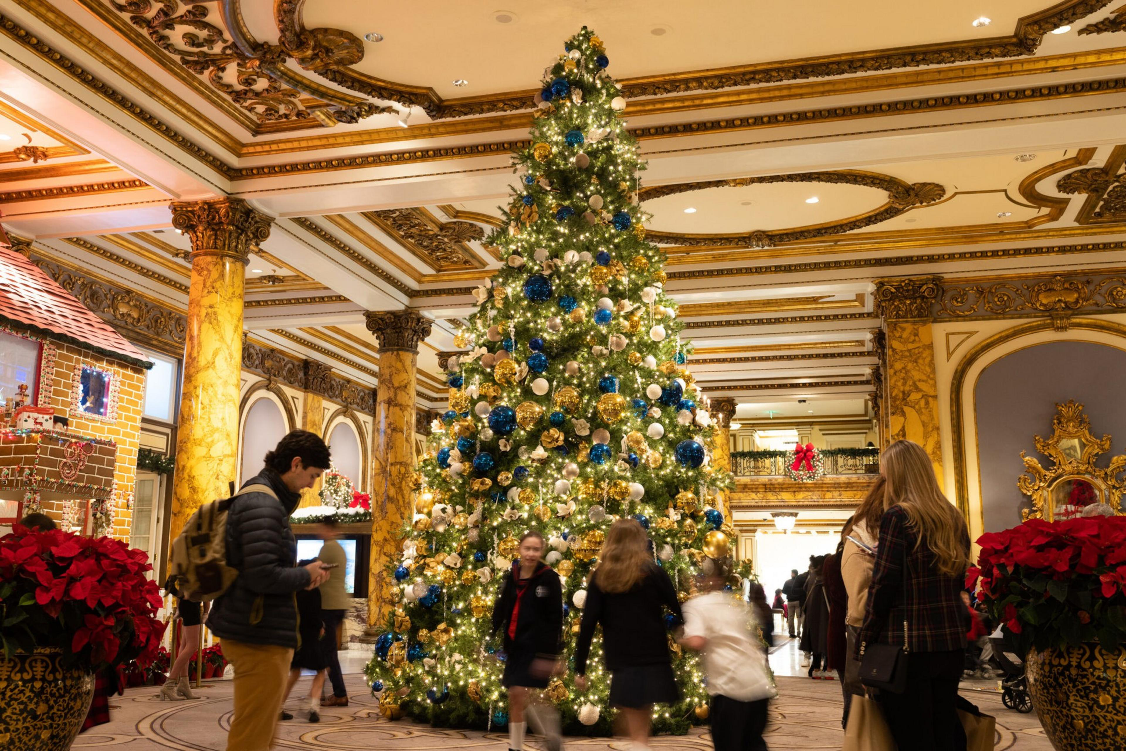 A large, decorated Christmas tree with blue, gold, and white ornaments stands in an ornate, marble-columned lobby with people admiring it.