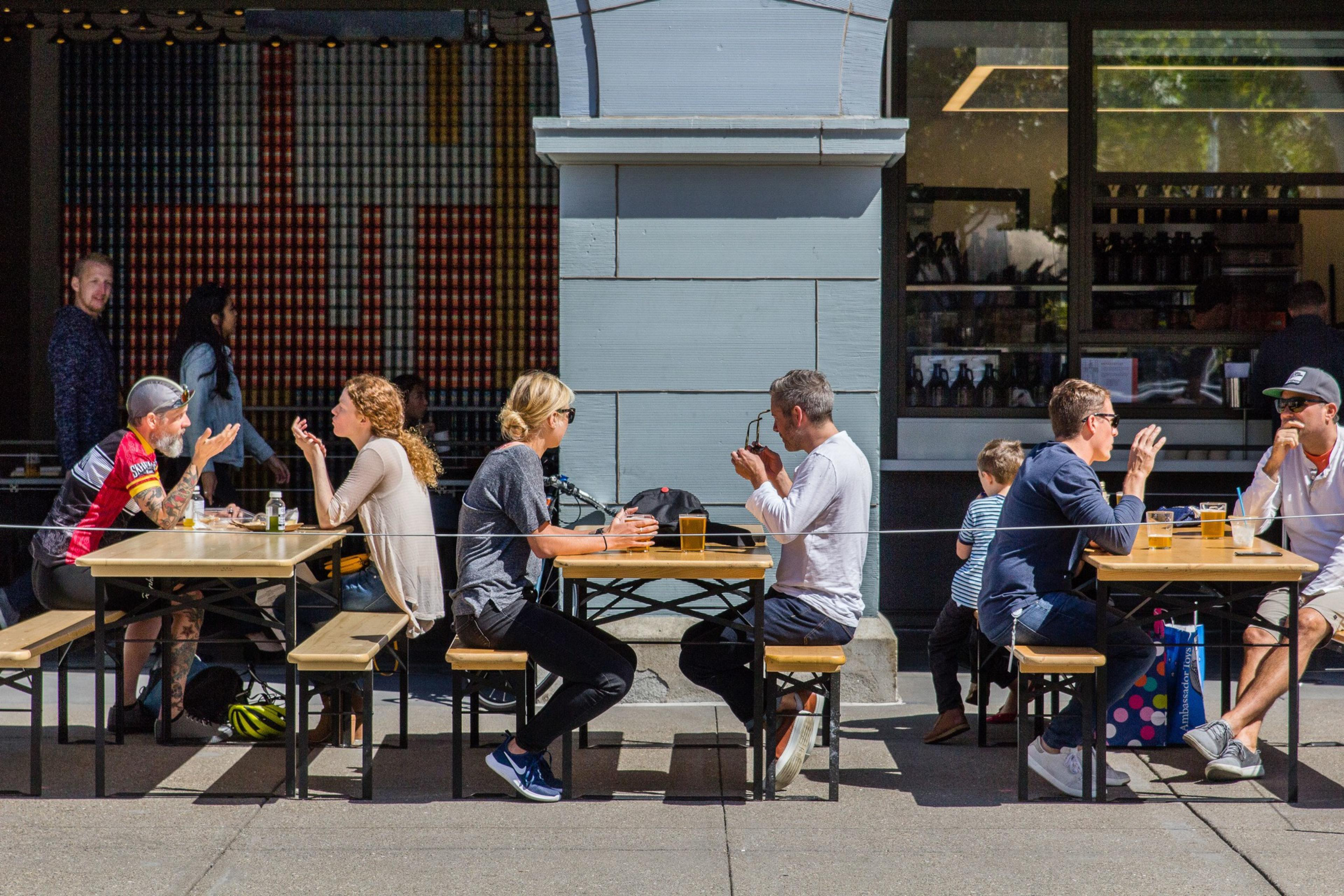 People sit at outdoor tables on a sunny day, chatting and enjoying drinks at a cafÃ©. A few individuals are seen walking behind them.