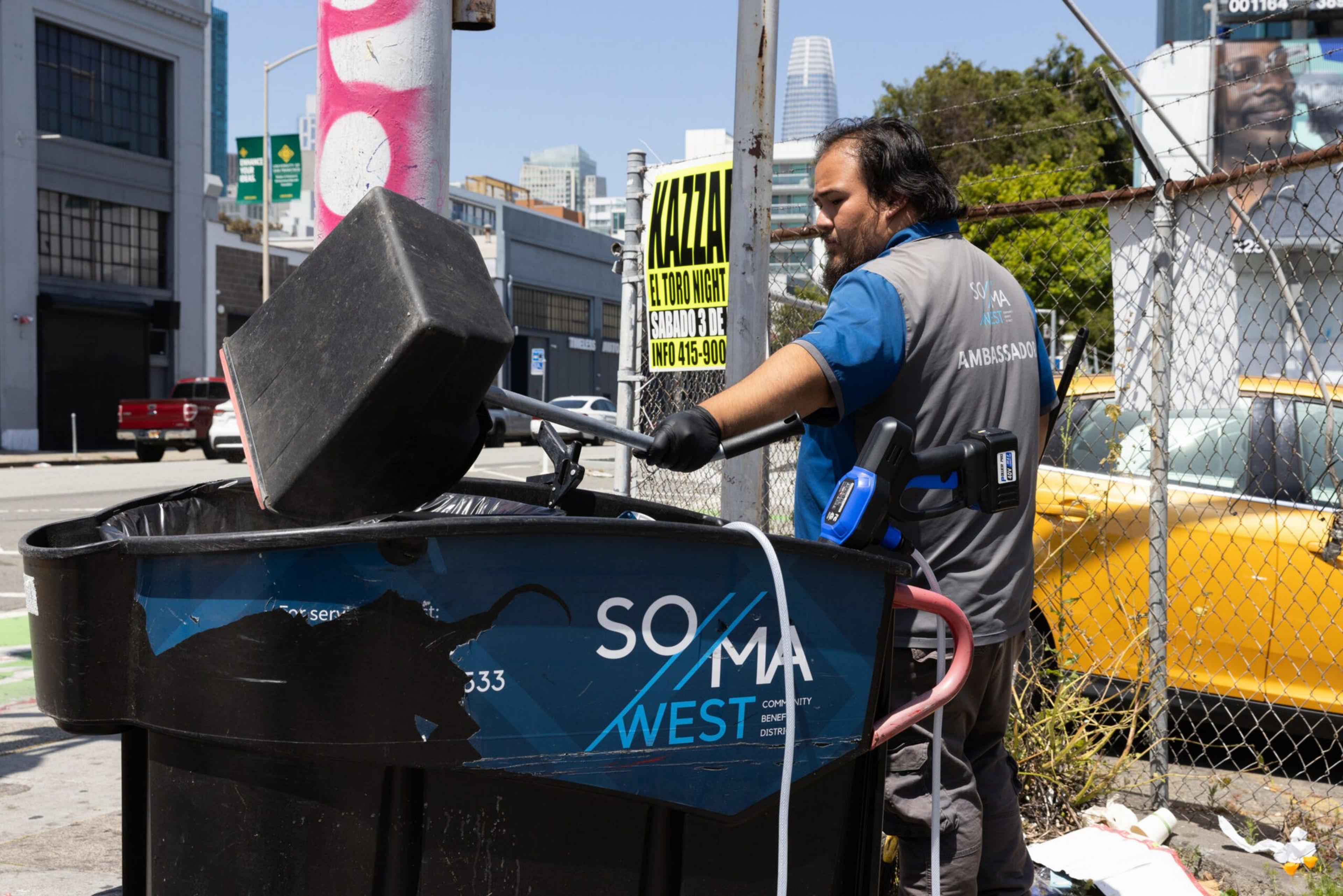 A sanitation worker wearing gloves and a vest labeled "AMBASSADOR" empties a trash bin into a large black dumpster on a sunny urban street.