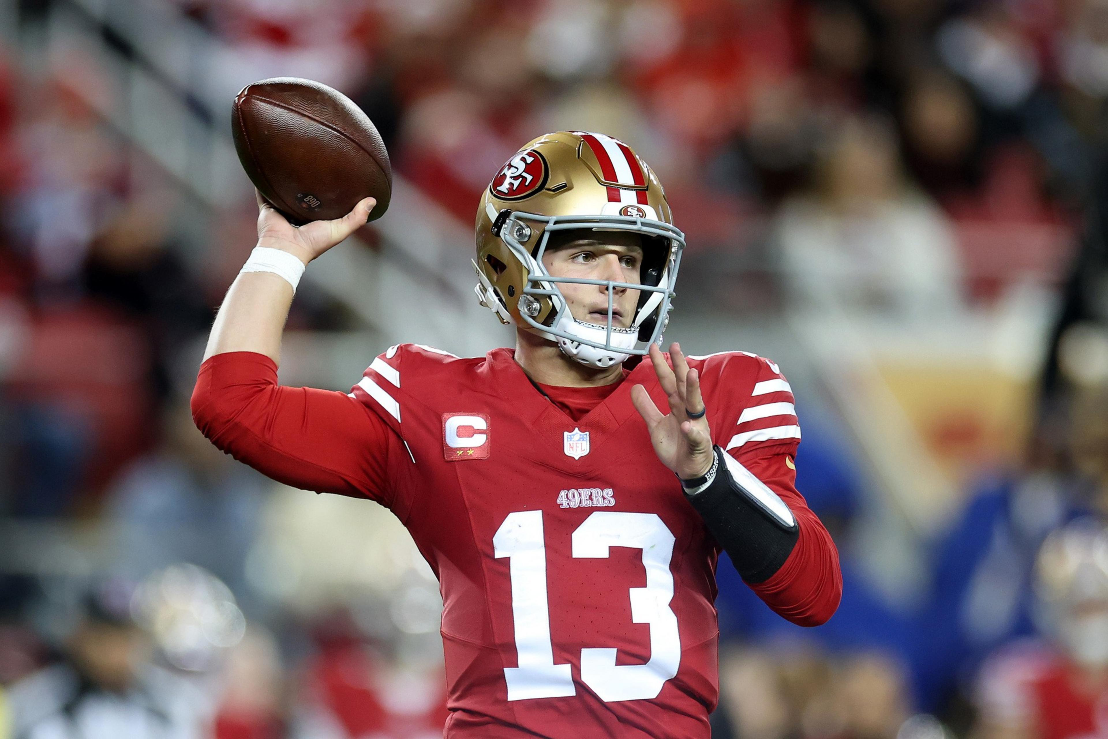 A football player in a red and gold uniform is poised to throw a football. He wears a helmet with a faceguard, and the setting suggests a professional game.