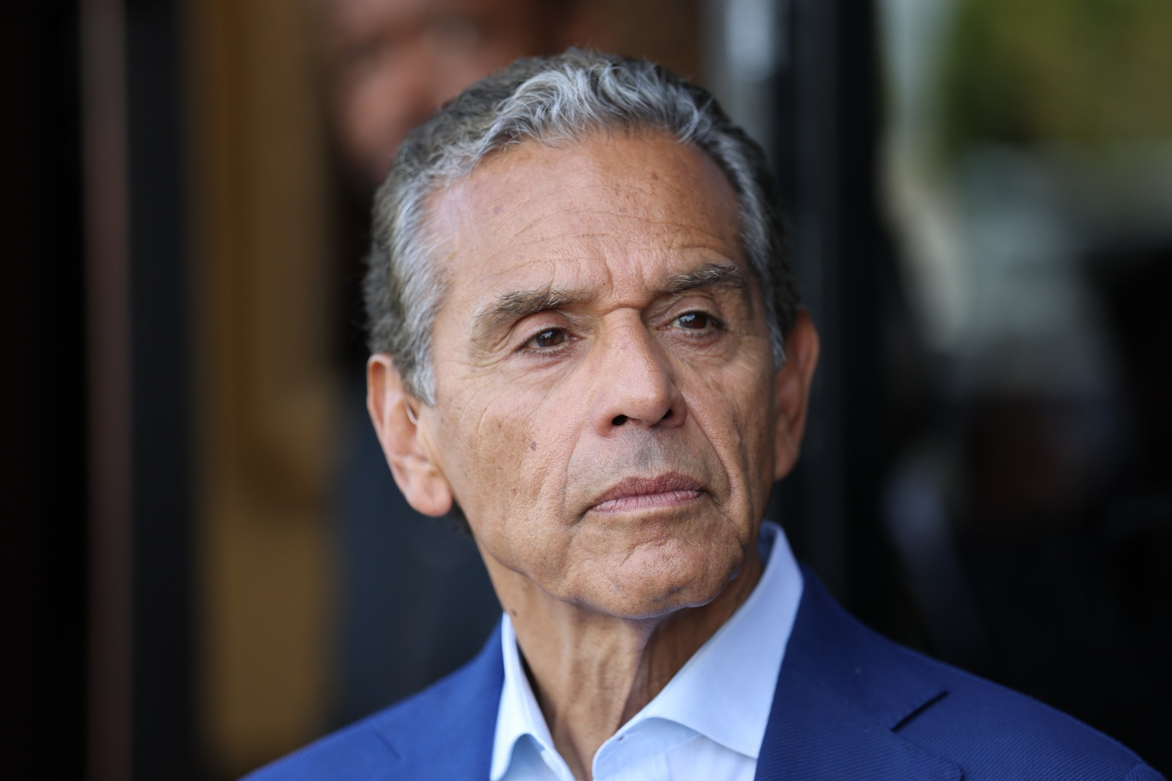 An older man with gray hair and a serious expression wears a blue suit jacket and white shirt, looking slightly to his left.