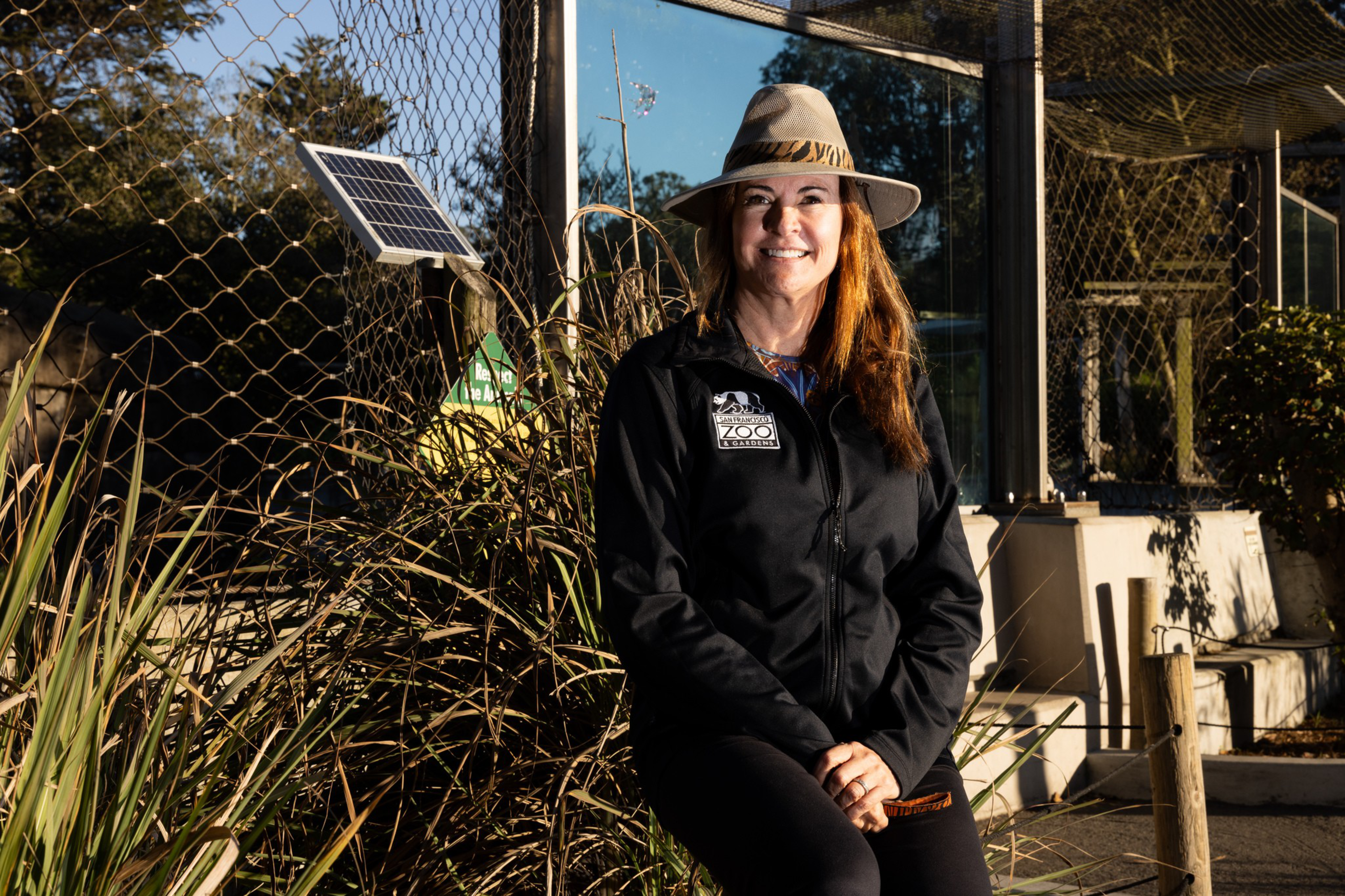 A person wearing a wide-brimmed hat and a black jacket with a zoo logo stands smiling beside a mesh enclosure and solar panel with foliage around.