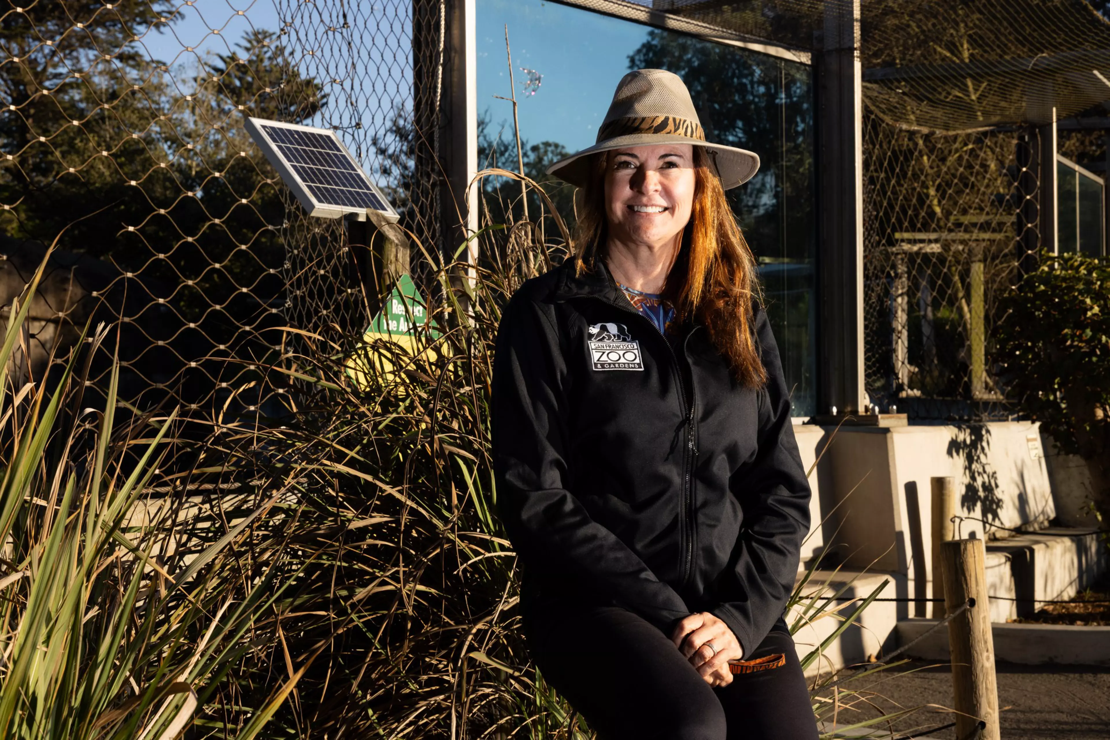 A person wearing a wide-brimmed hat and a black jacket with a zoo logo stands smiling beside a mesh enclosure and solar panel with foliage around.