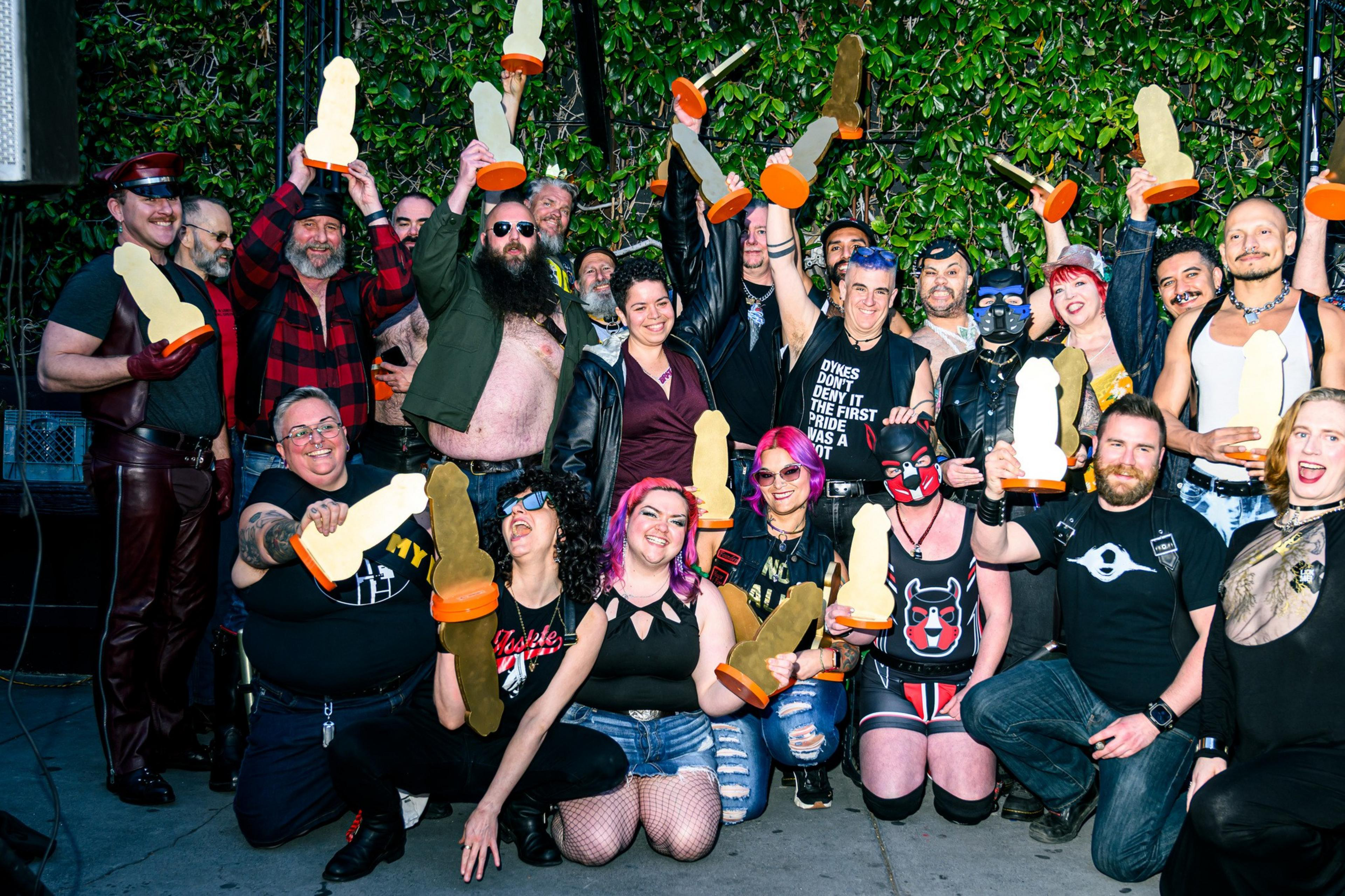 A group of people is gathered outdoors, many holding yellow trophies with orange bases. They appear joyful, dressed in various styles, and are standing against greenery.