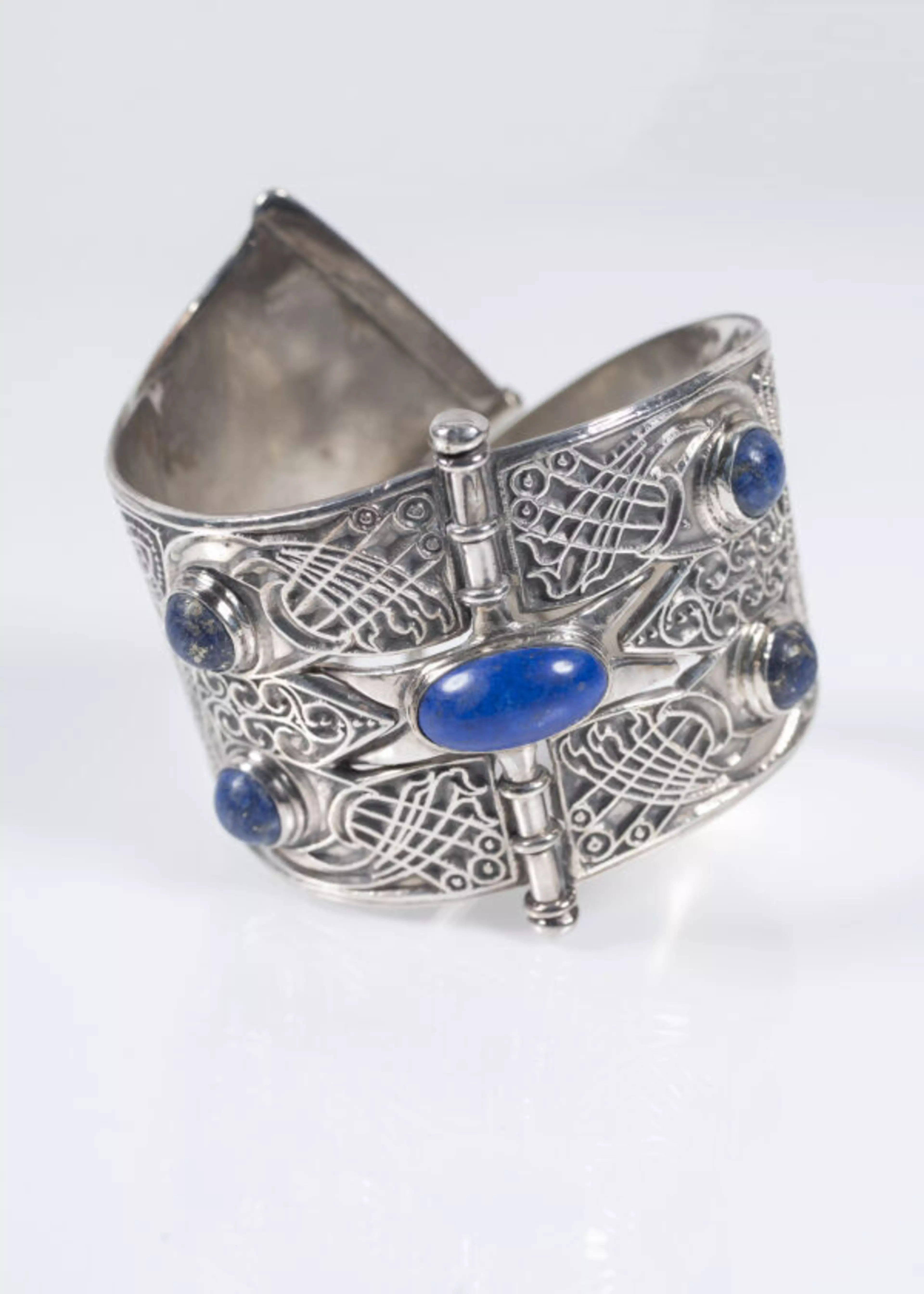 A wide silver cuff bracelet features intricate filigree designs with five oval blue stones set symmetrically around a central horizontal bar.