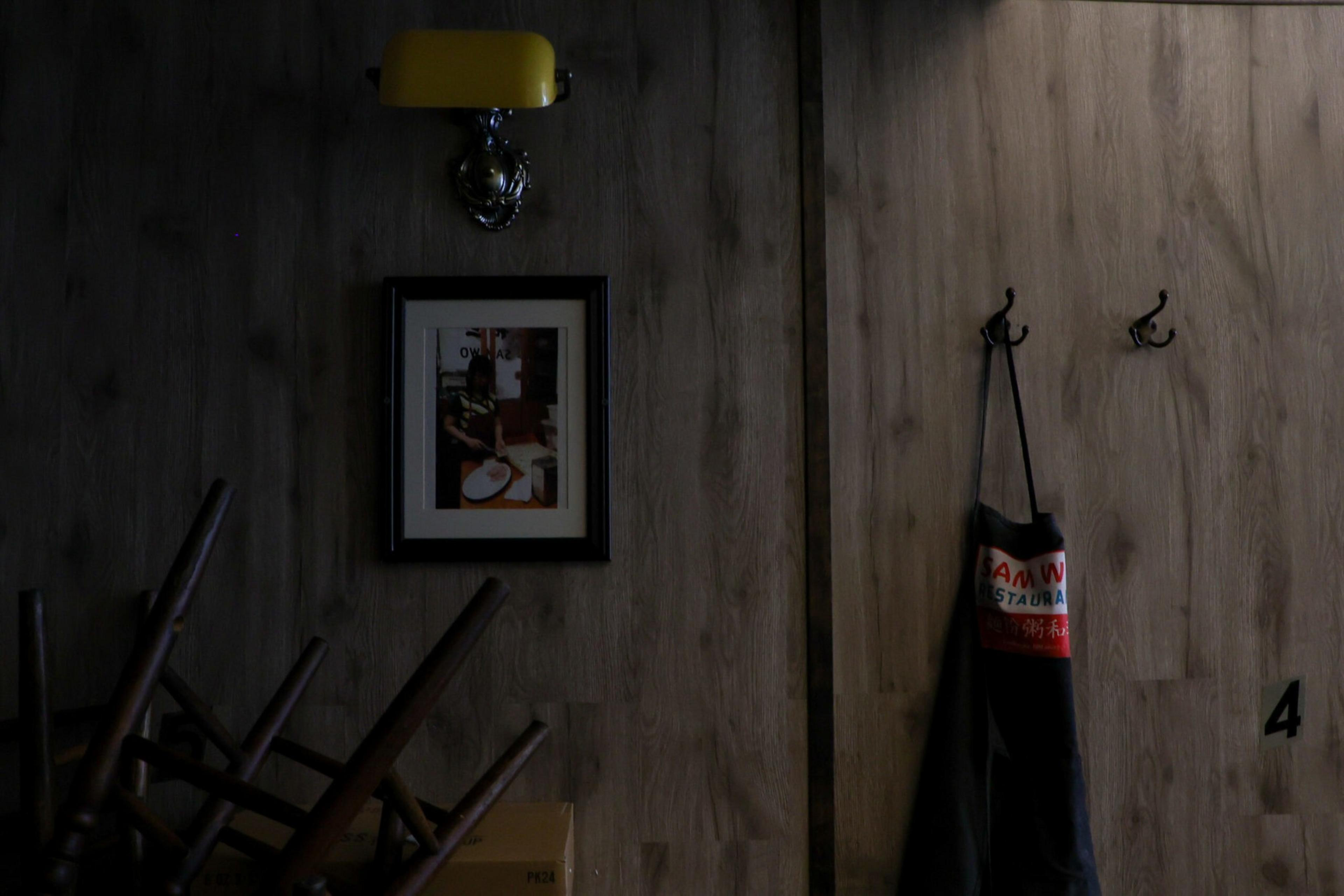 A wooden wall with a framed photo, a yellow wall lamp above it, three chair legs visible on the left, and a hanging black apron on the right.