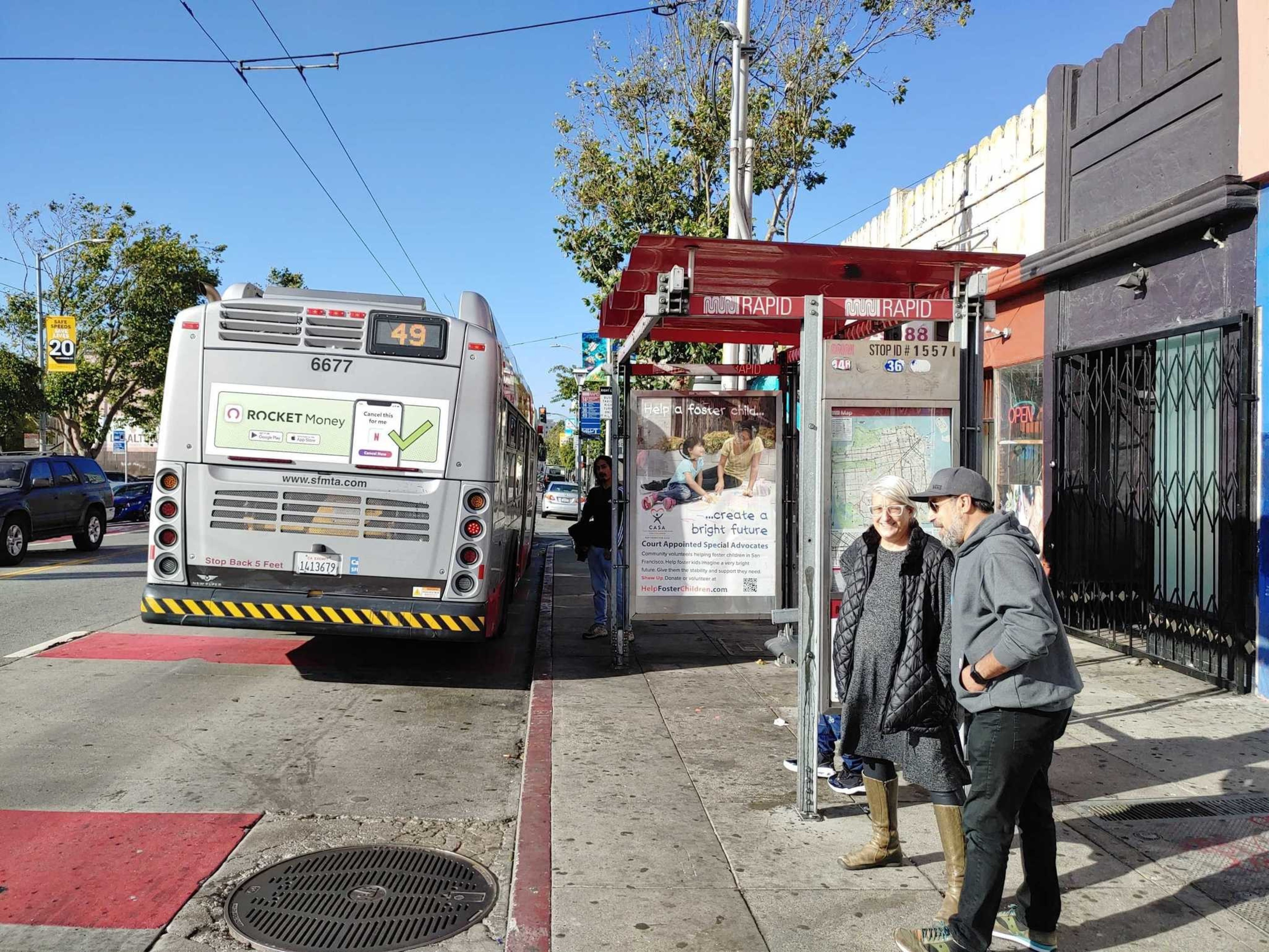 A bus numbered 49 is stopped at a bus stop where two people are standing and talking near the shelter, with a sidewalk and trees in the background.