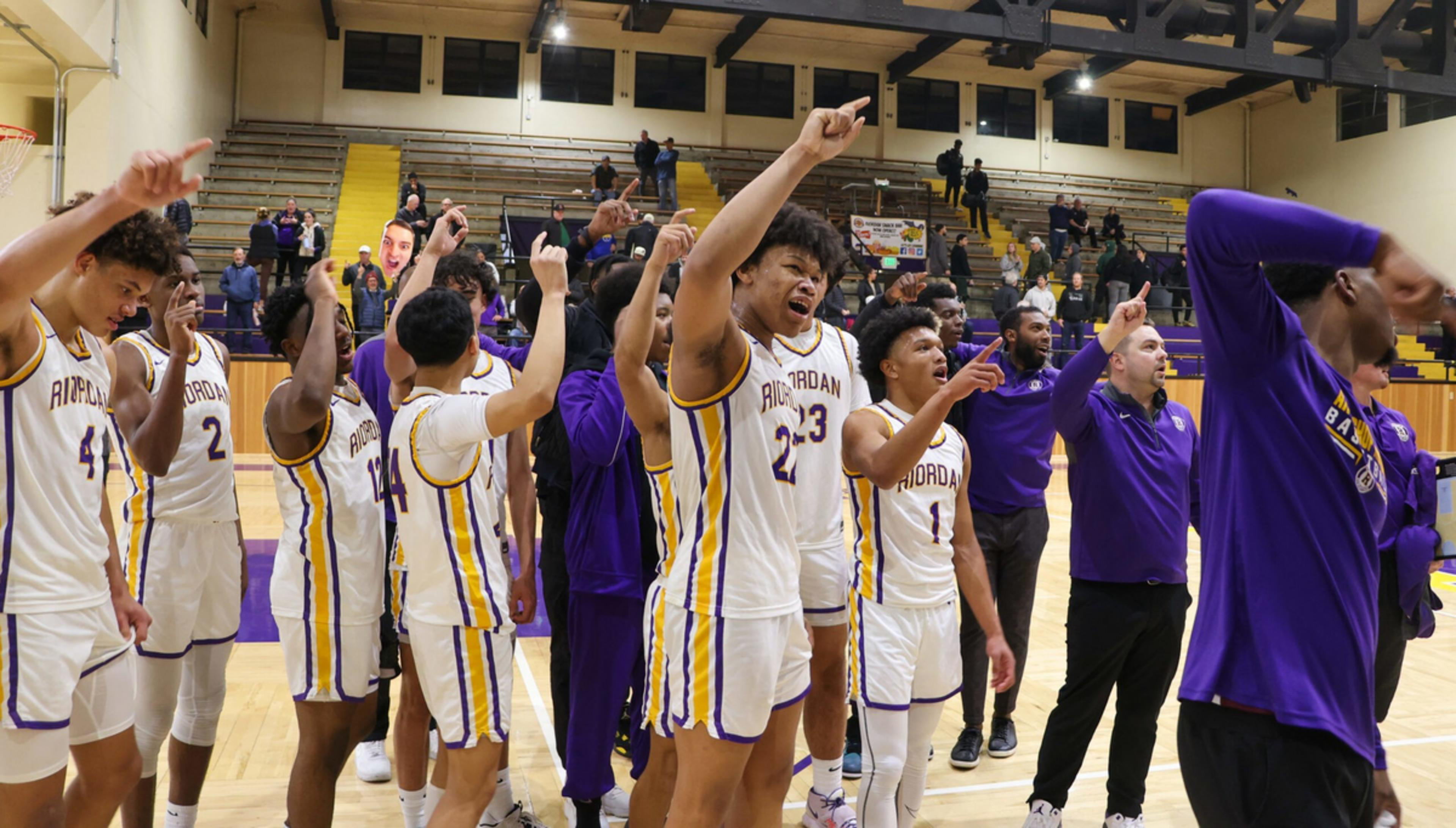 A basketball team in white and purple uniforms stands on the court celebrating with raised arms and enthusiastic expressions.