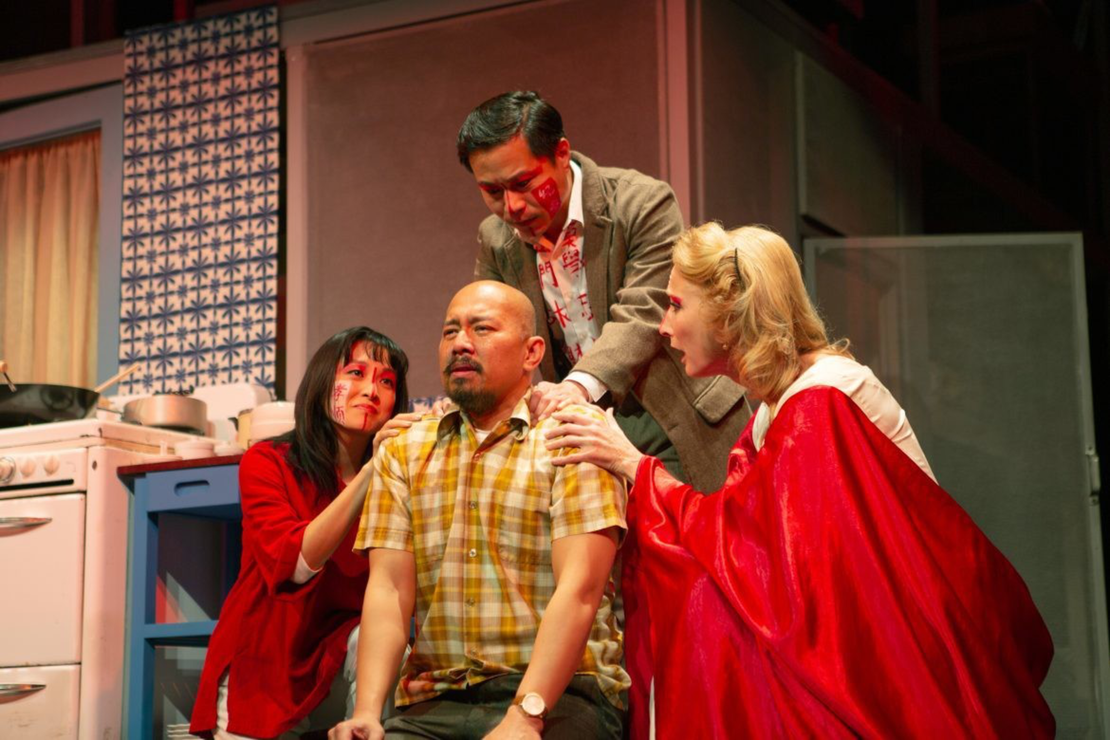 Three people gently console a man in a yellow plaid shirt who looks emotional, while two of them have red markings on their faces and one wears a red cape.