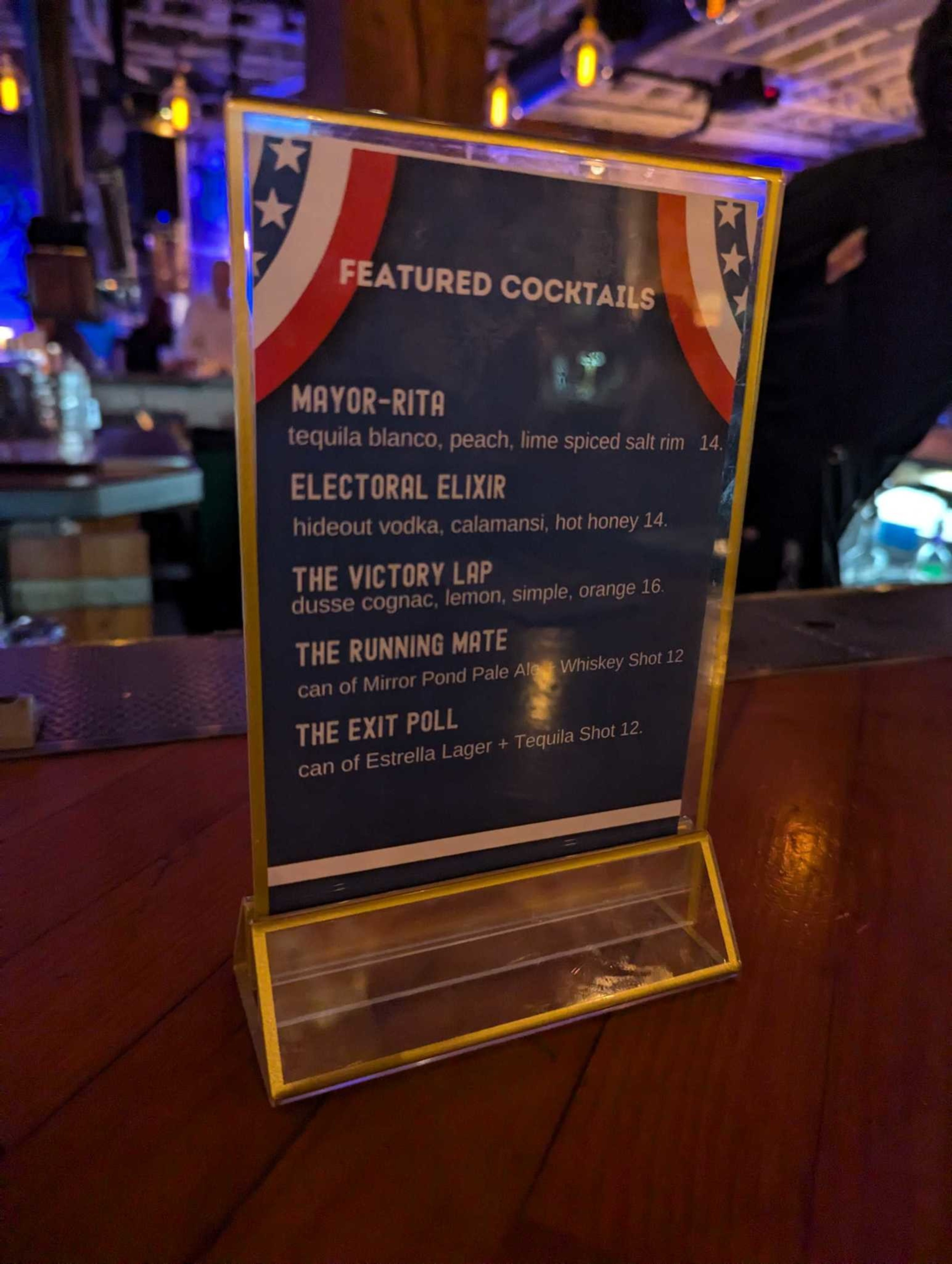 The image shows a menu with a list of featured cocktails, each with humorous election-themed names, displayed in a dimly lit bar setting.