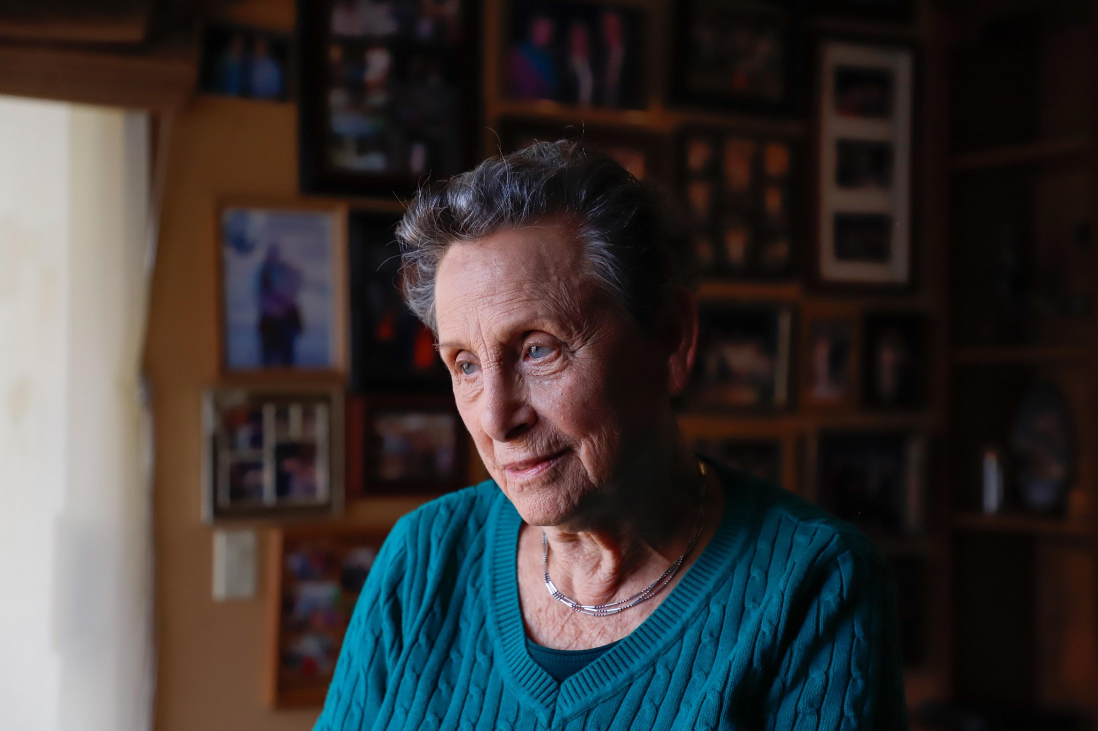 An elderly woman with short gray hair and a teal sweater looks thoughtfully toward a softly lit window, with a photo wall blurred behind her.
