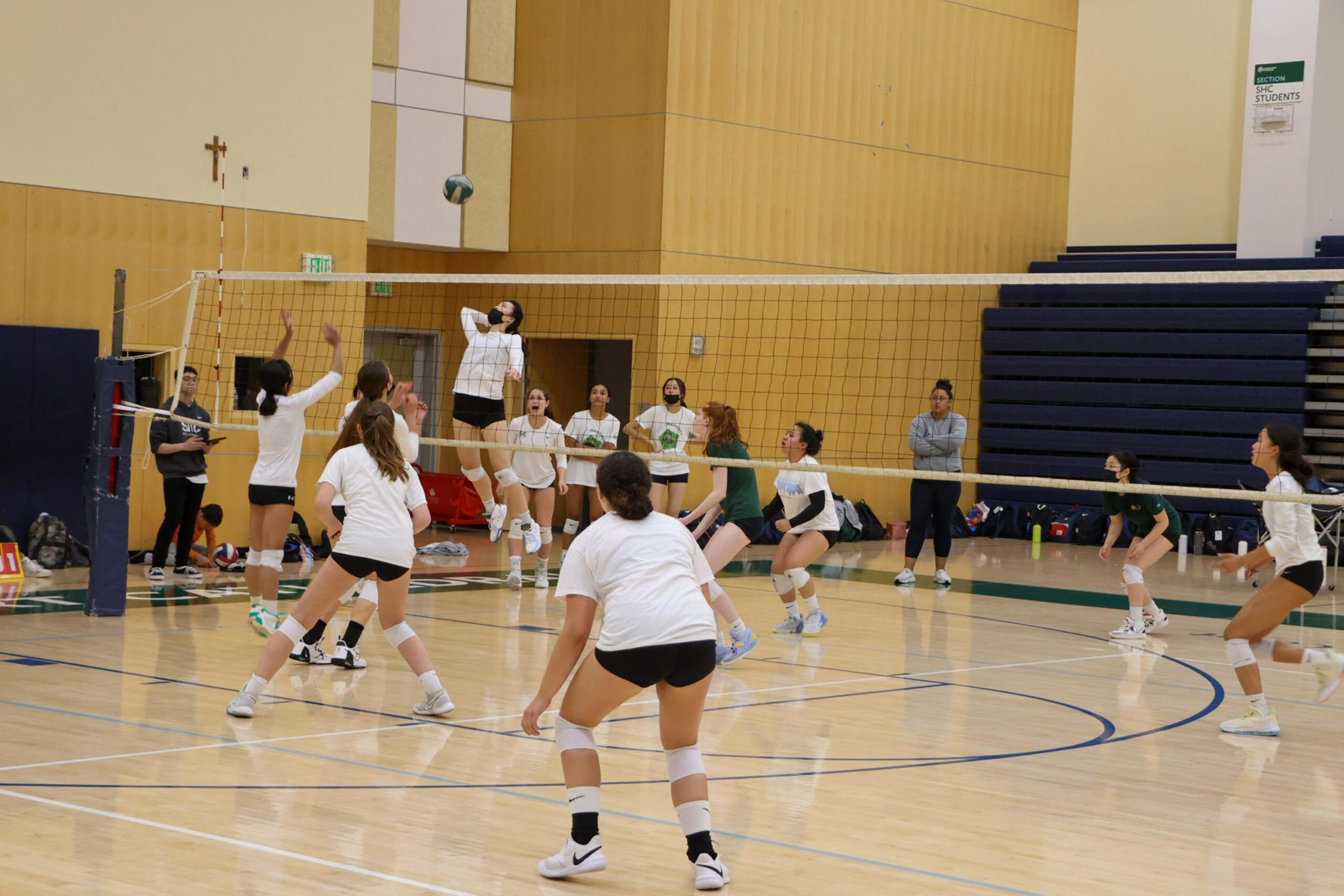A women's volleyball game is in progress in a gym, with players in white and green uniforms actively engaged near the net and others ready in position.