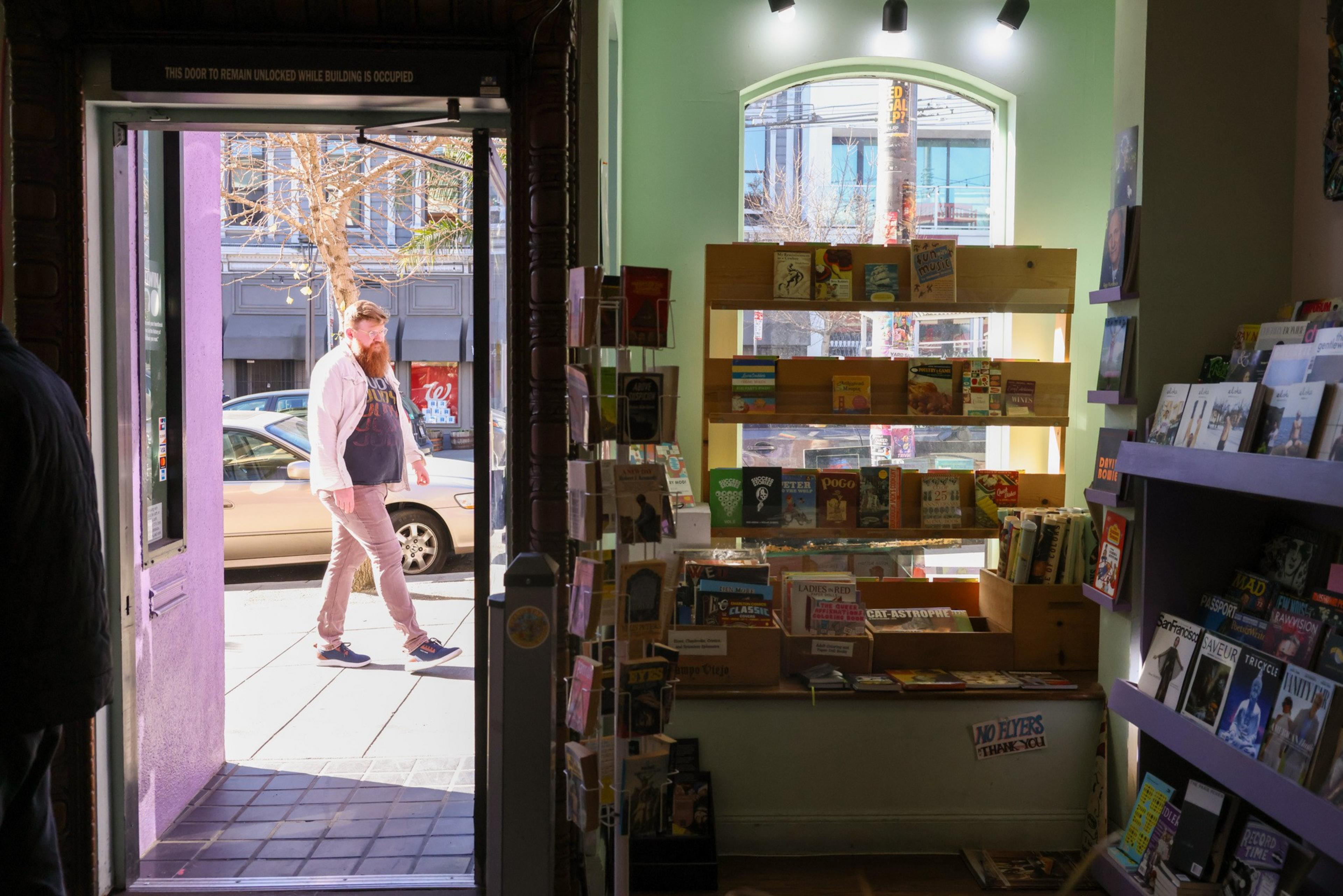 A man walks past an open bookstore door, with a display of books and magazines visible inside. Sunlight streams in, casting shadows on the floor.