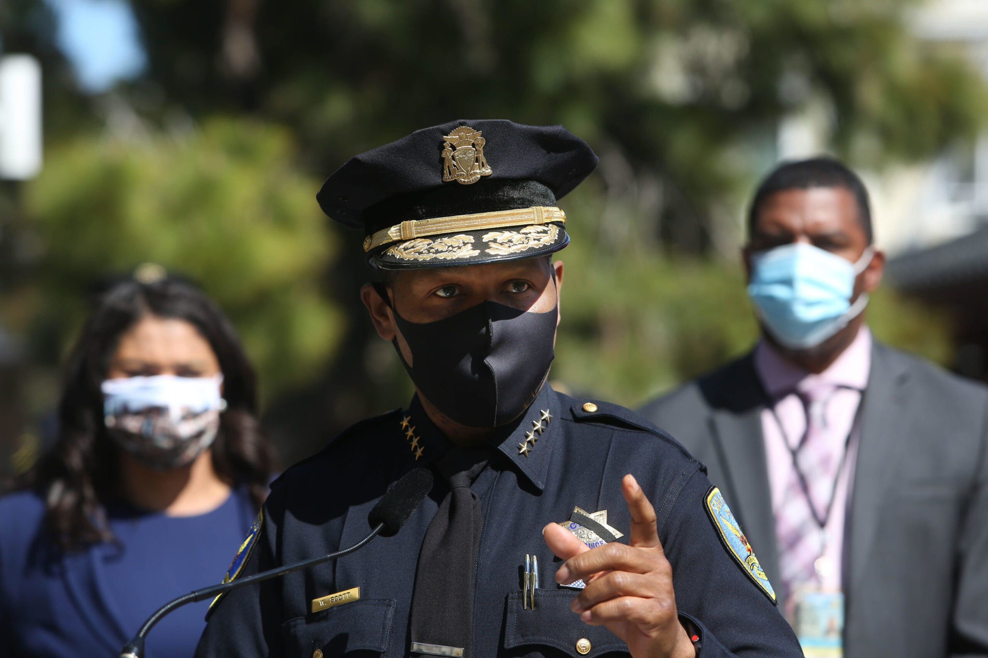 A police officer in uniform and a black face mask speaks, gesturing with his hand, while two masked individuals stand behind him outdoors.