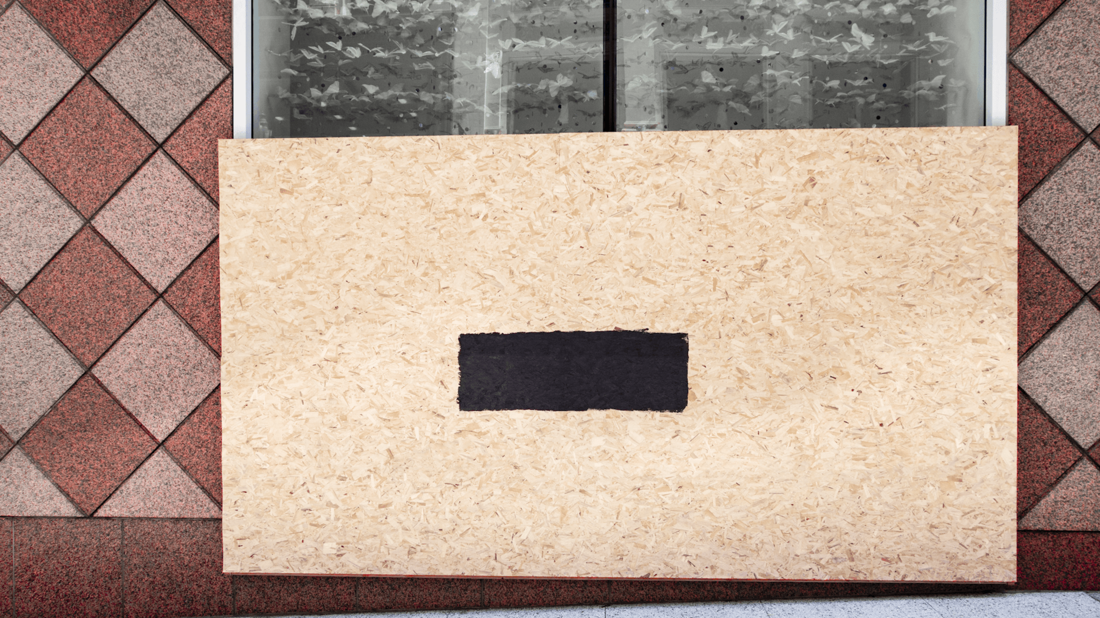 A boarded-up window with a rectangular black patch in the center is set against a red and gray diamond-patterned wall.