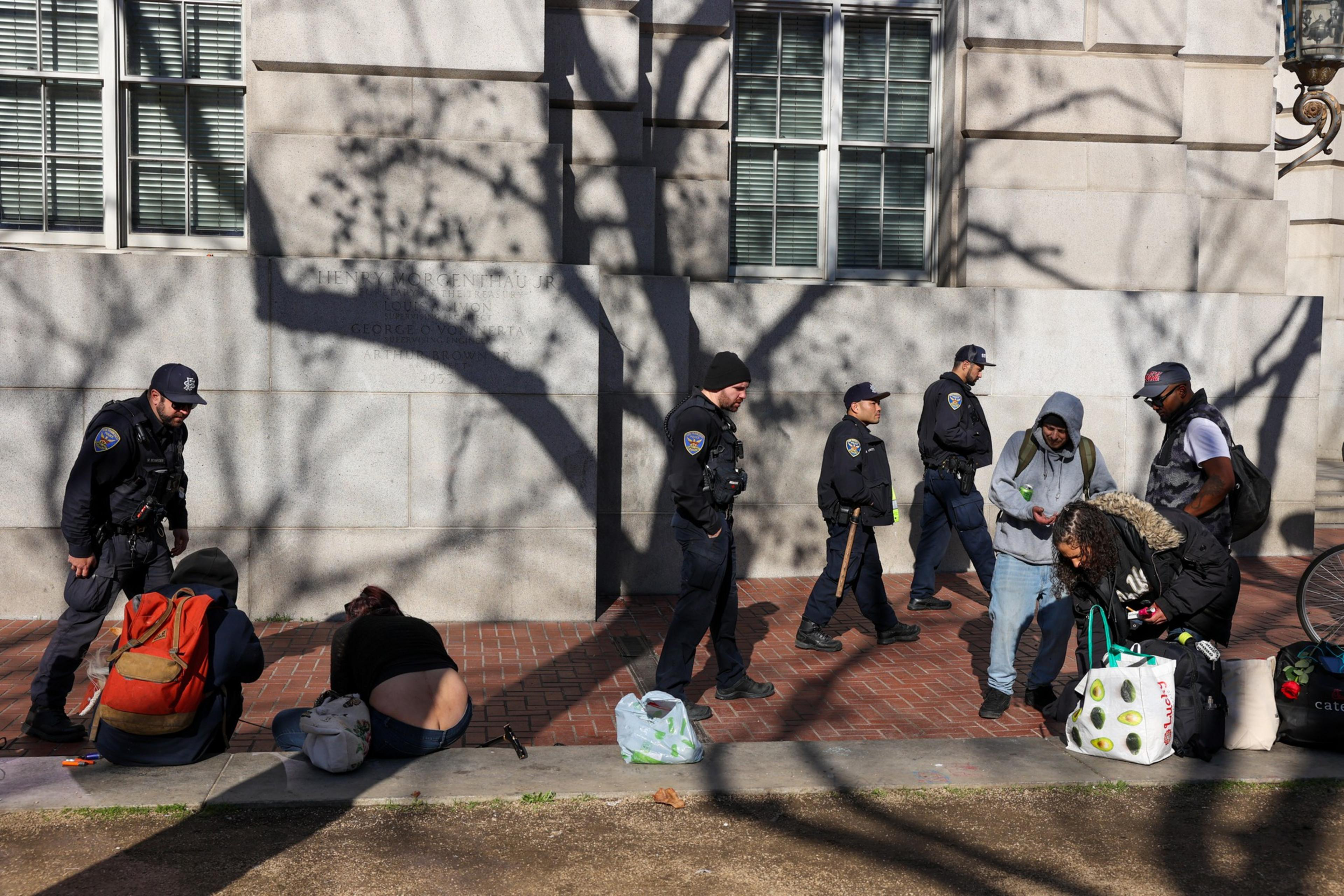 Four police officers walk along a brick sidewalk while five individuals with bags sit or stand near a building casting tree shadows.