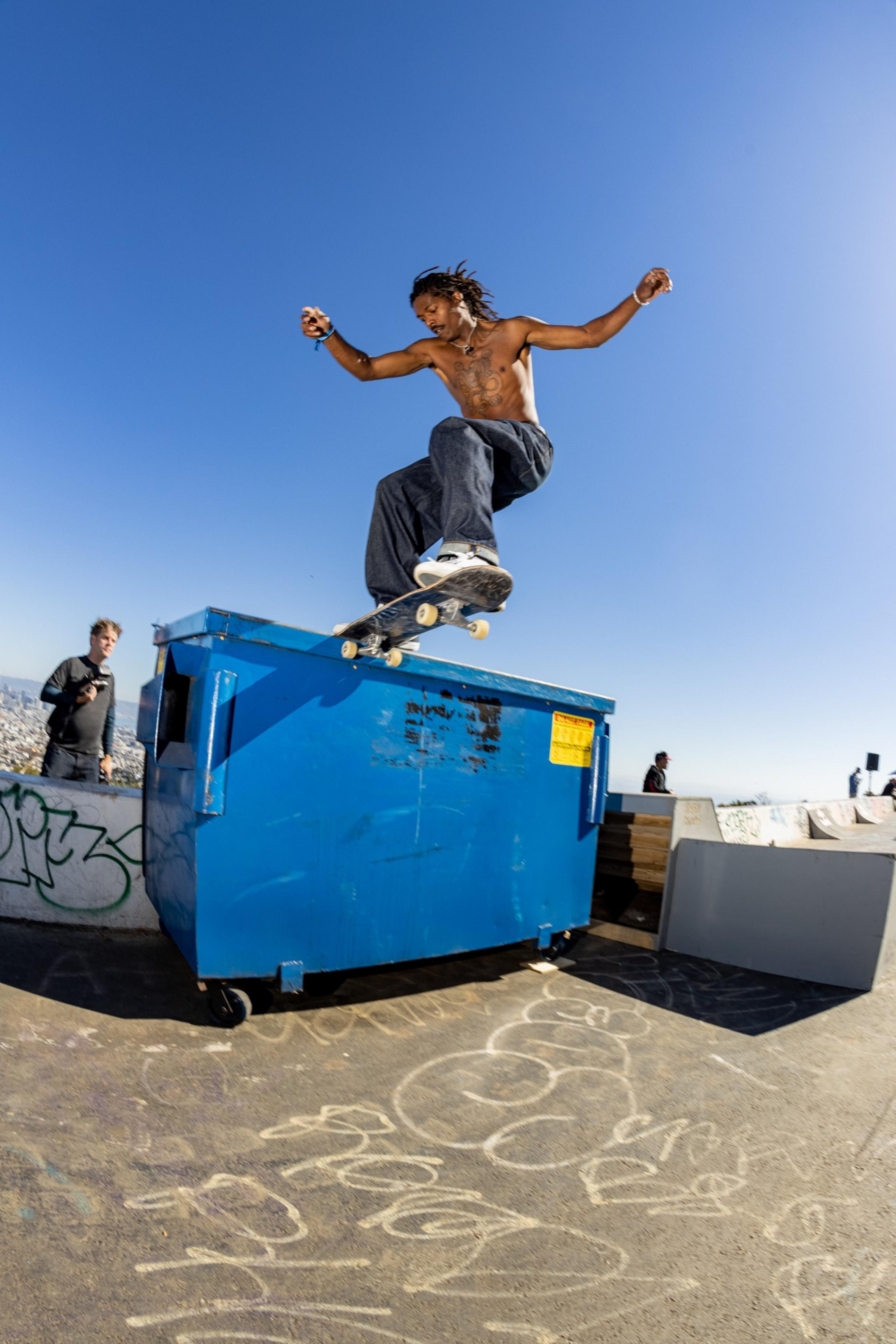 A shirtless skateboarder performs a trick off a blue dumpster under a clear sky. A person in the background watches, and graffiti covers the ground and walls.