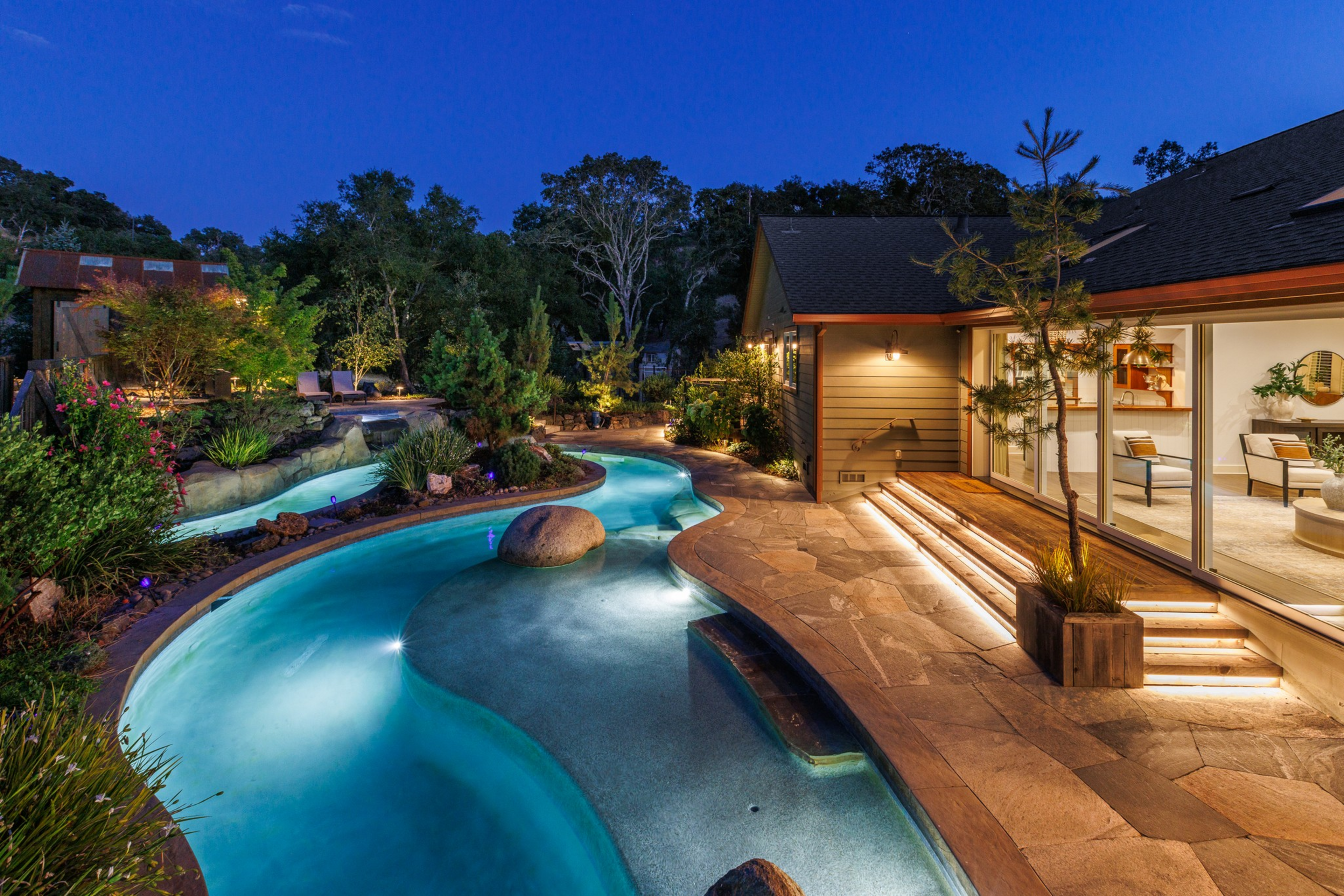 A curving, illuminated pool winds through a landscaped backyard beside a house with large glass windows and lighted stone stairs.
