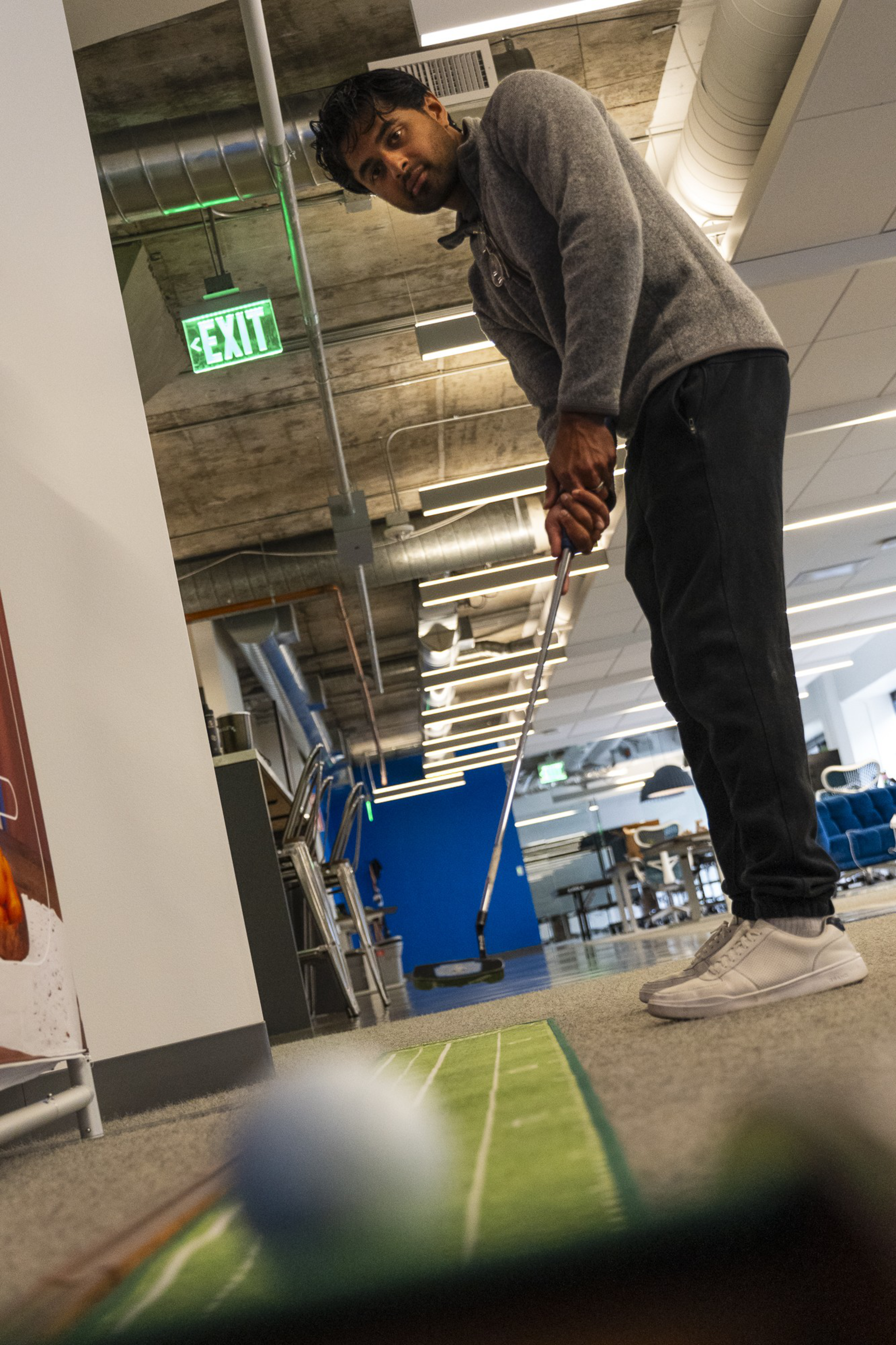 A person in a gray sweater is putting a golf ball on an indoor mat in an office with industrial ceilings, a green EXIT sign, and blue accents.