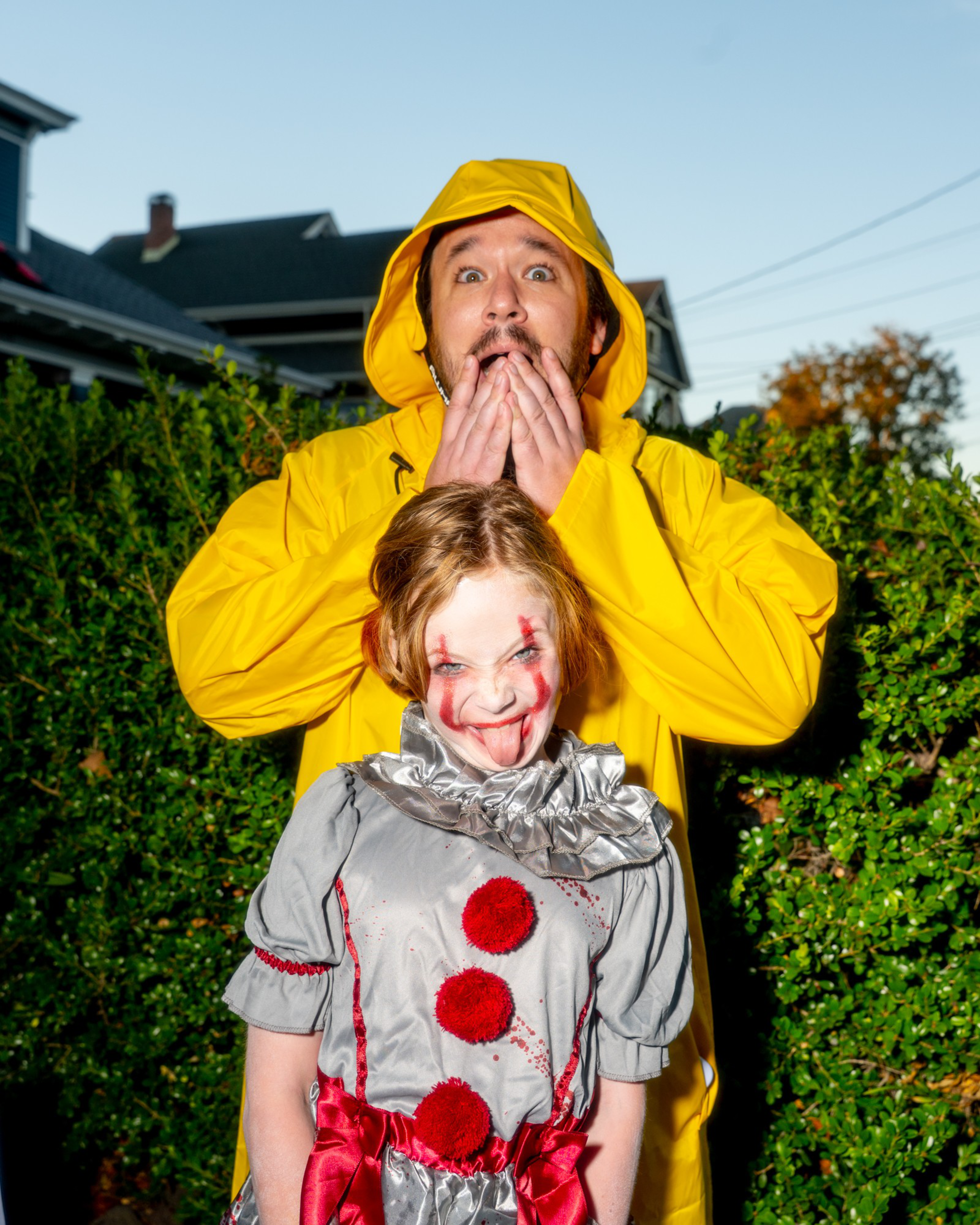 A man in a yellow raincoat looks shocked, covering his mouth, while a child in a bloody clown costume sticks out their tongue mischievously.