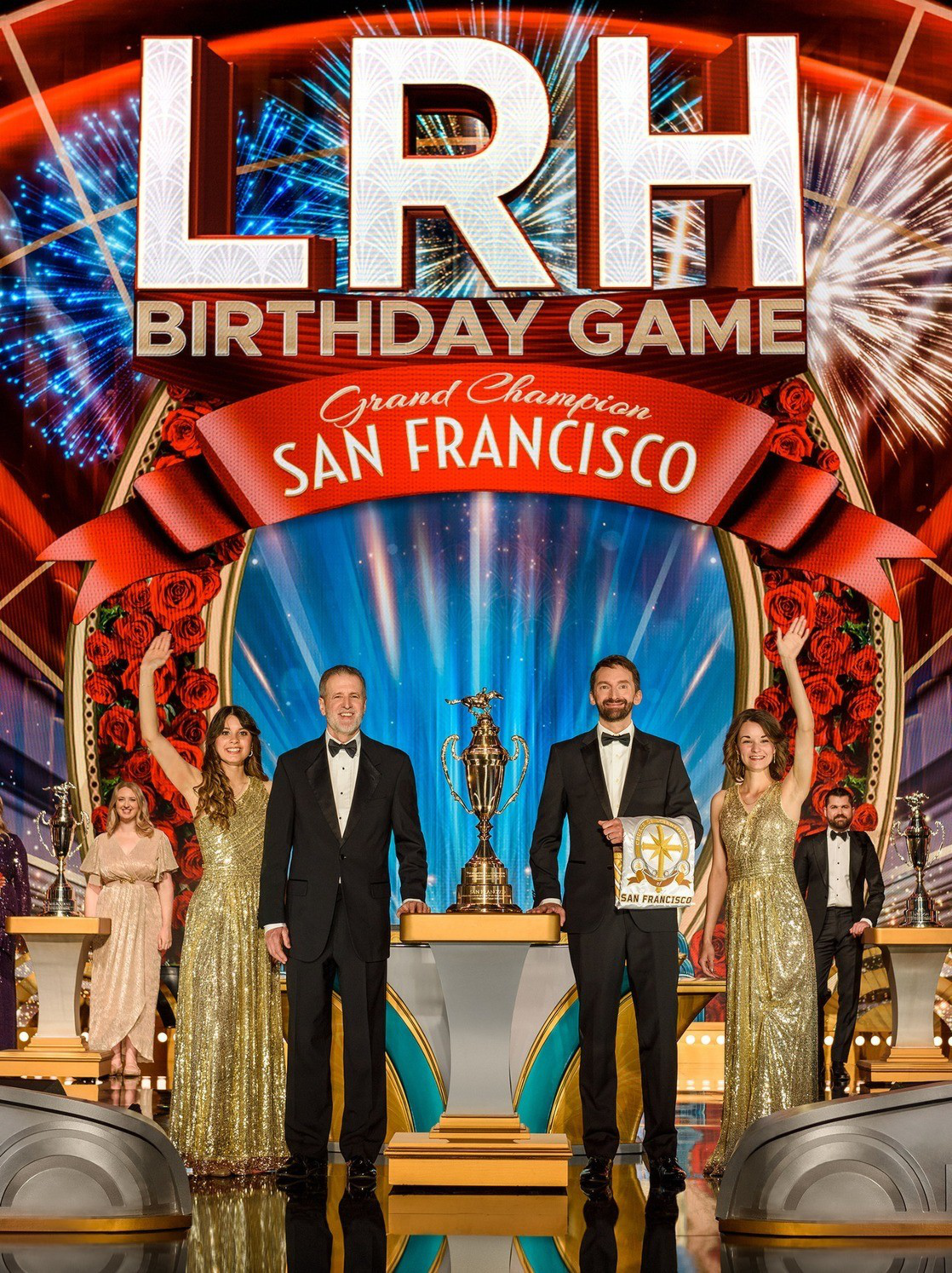 A group of smiling people in formal attire stand on a stage with a large trophy and a plaque reading &quot;San Francisco.&quot; The backdrop features the text &quot;LRH Birthday Game.&quot;