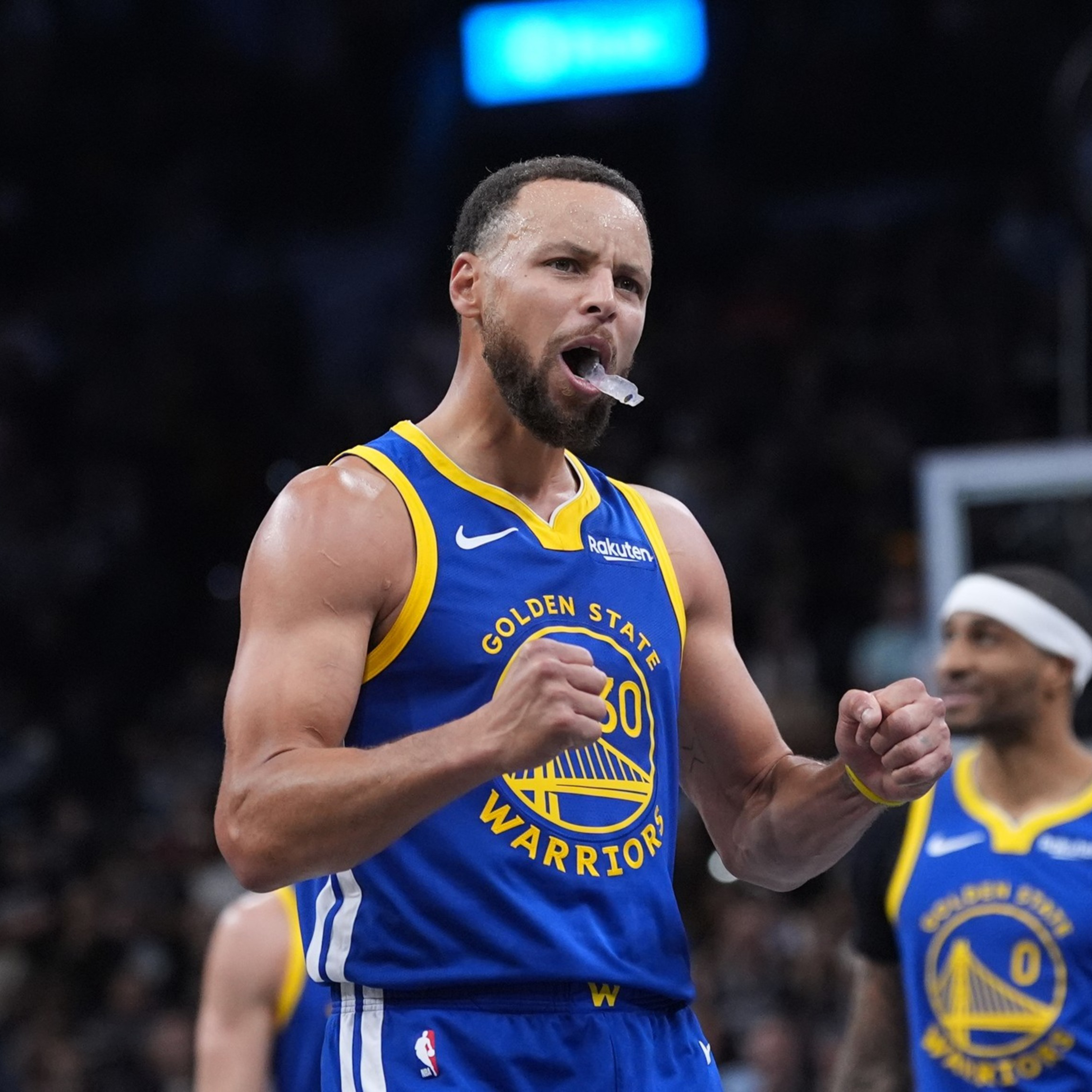 A Golden State Warriors player clenches his fists and cheers with a mouthguard in his mouth, while a teammate smiles in the background near the basketball hoop.