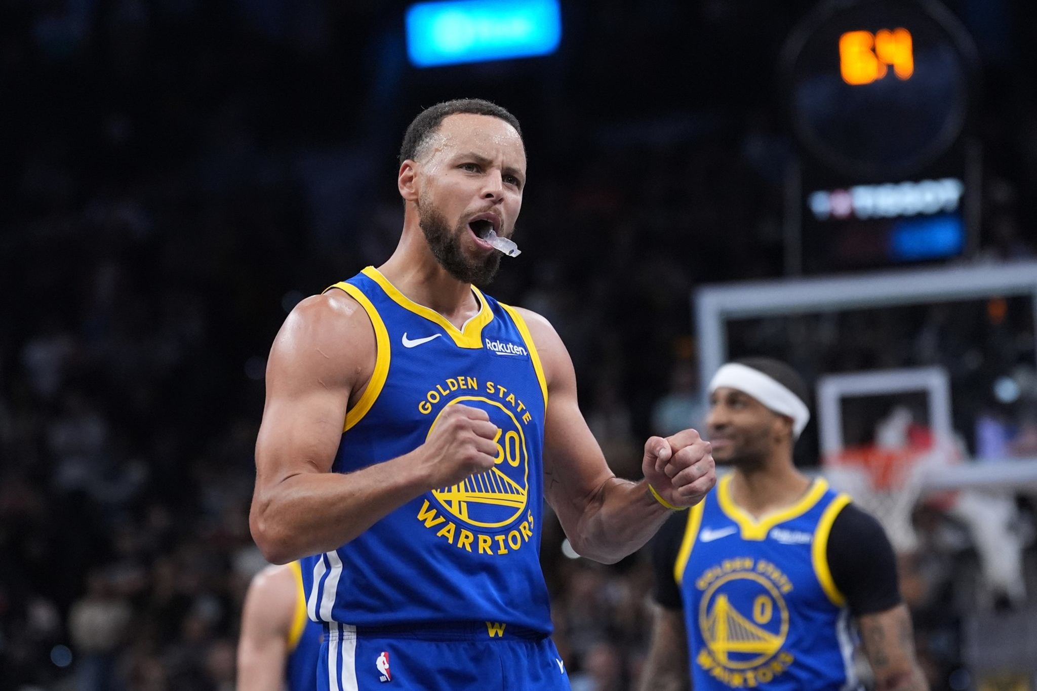 A Golden State Warriors player clenches his fists and cheers with a mouthguard in his mouth, while a teammate smiles in the background near the basketball hoop.