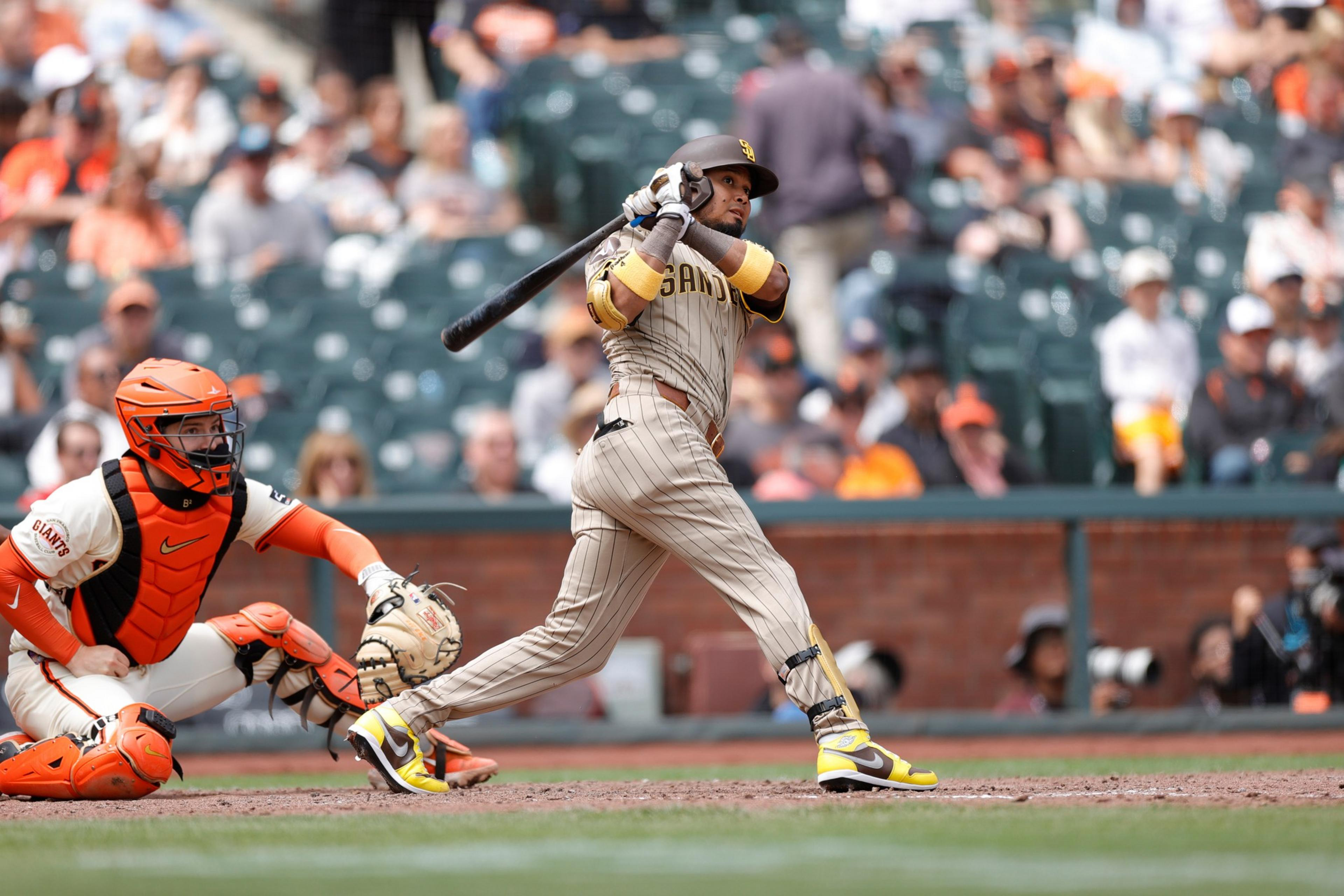 A baseball player in gray and yellow swings a bat as a catcher in orange gear crouches behind home plate, with blurred fans in the stadium seats behind them.