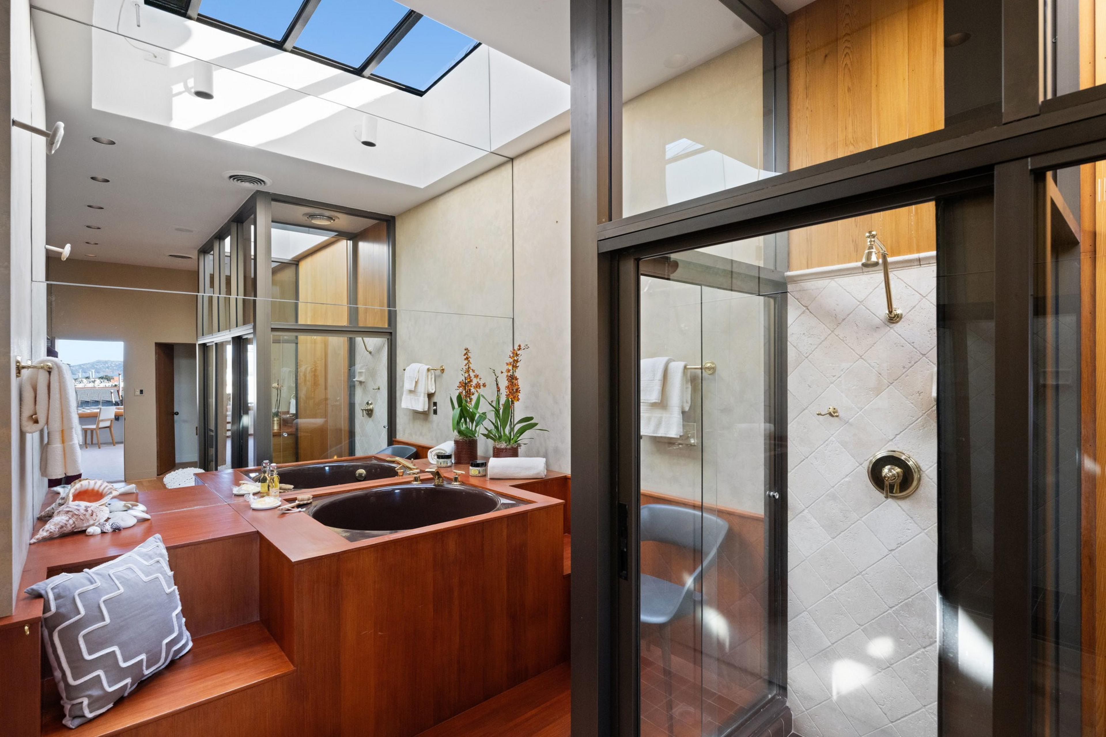 The image shows a modern bathroom with a skylight, wooden accents, a black bathtub, glass shower, and plants. Natural light illuminates the room.